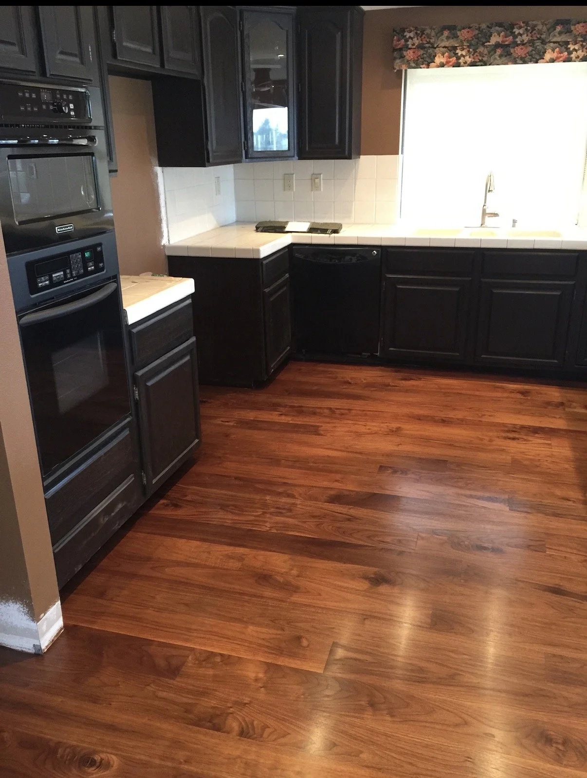 Kitchen with dark cabinets, white tiled countertop, wooden floor, and a window above the sink.