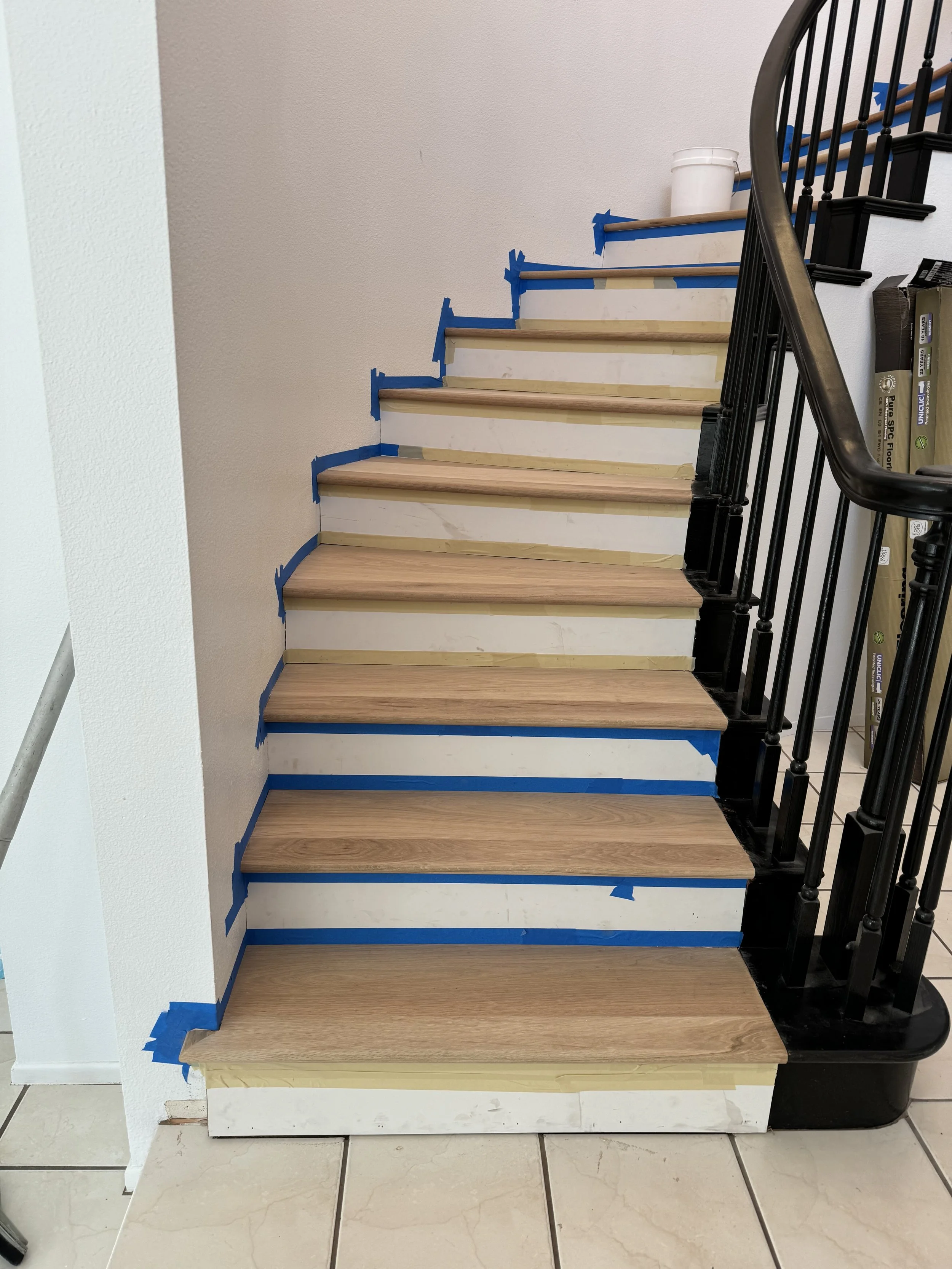 Staircase under renovation with unfinished wooden steps, blue painter's tape on edges, and a black metal railing. A white container is on the upper landing, and construction materials are visible.