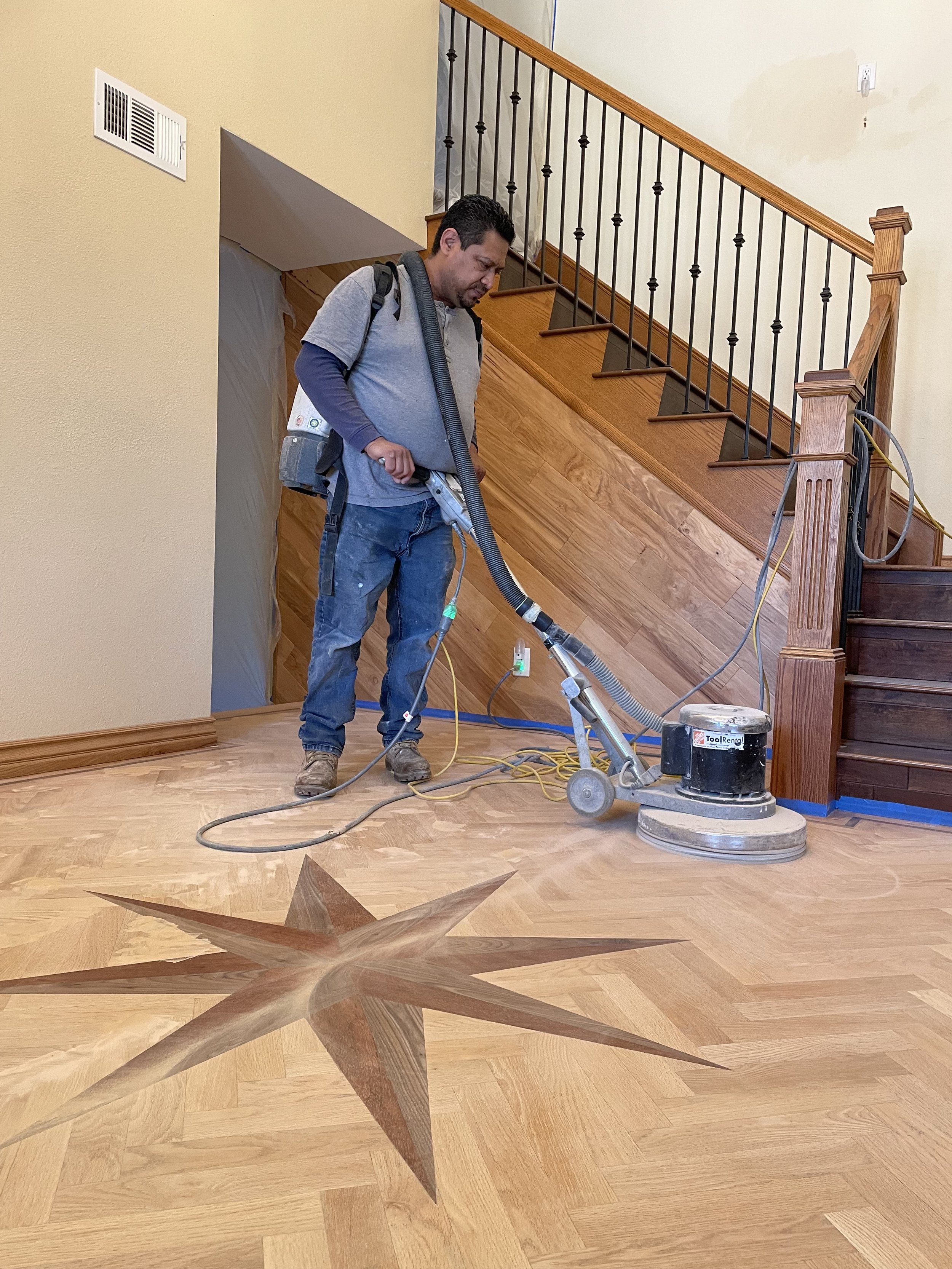 A man using a floor sander on a hardwood floor in a residential setting near a staircase.