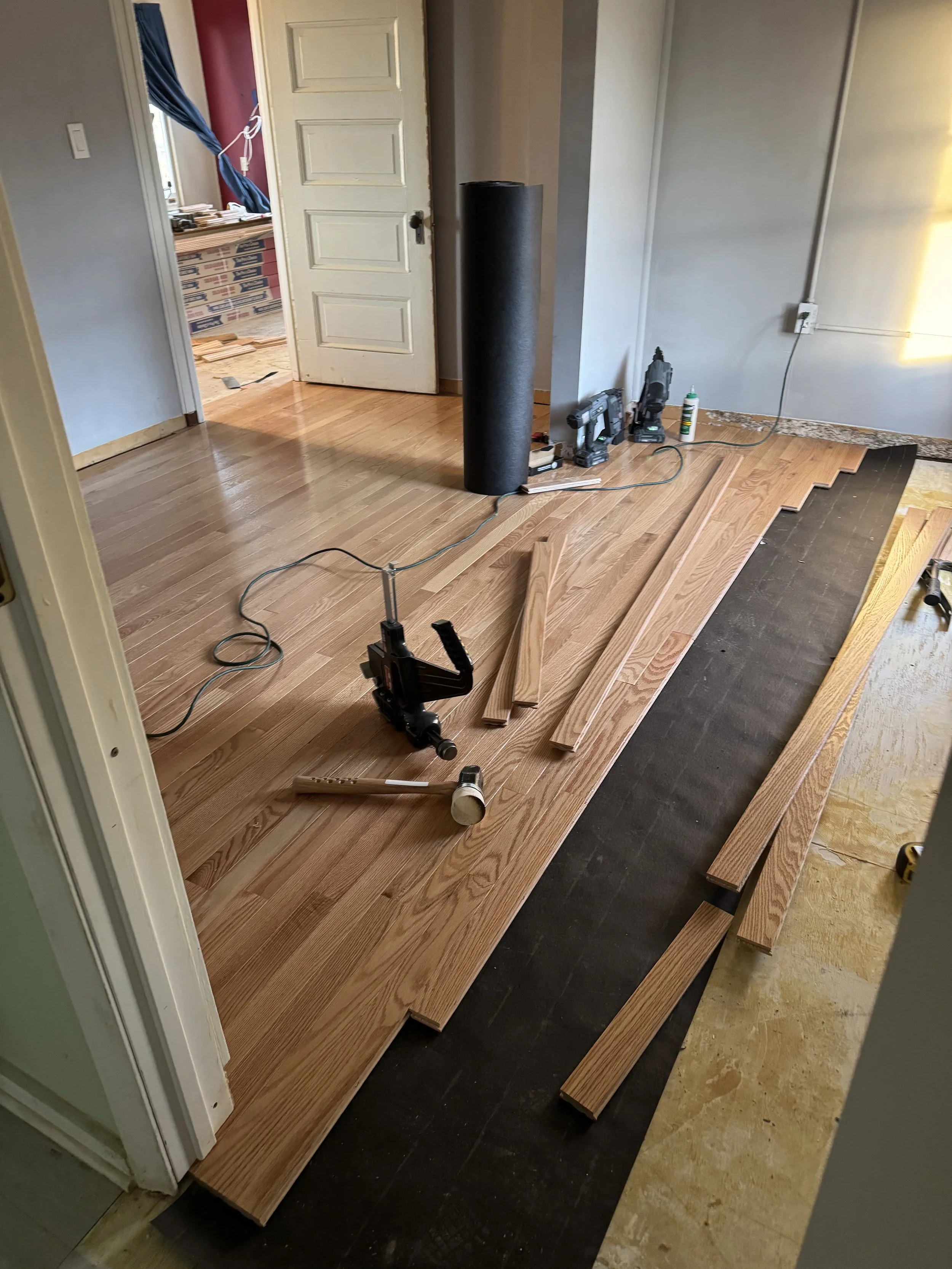 Room under renovation with hardwood flooring installation in progress, various tools and wooden planks scattered on the floor.