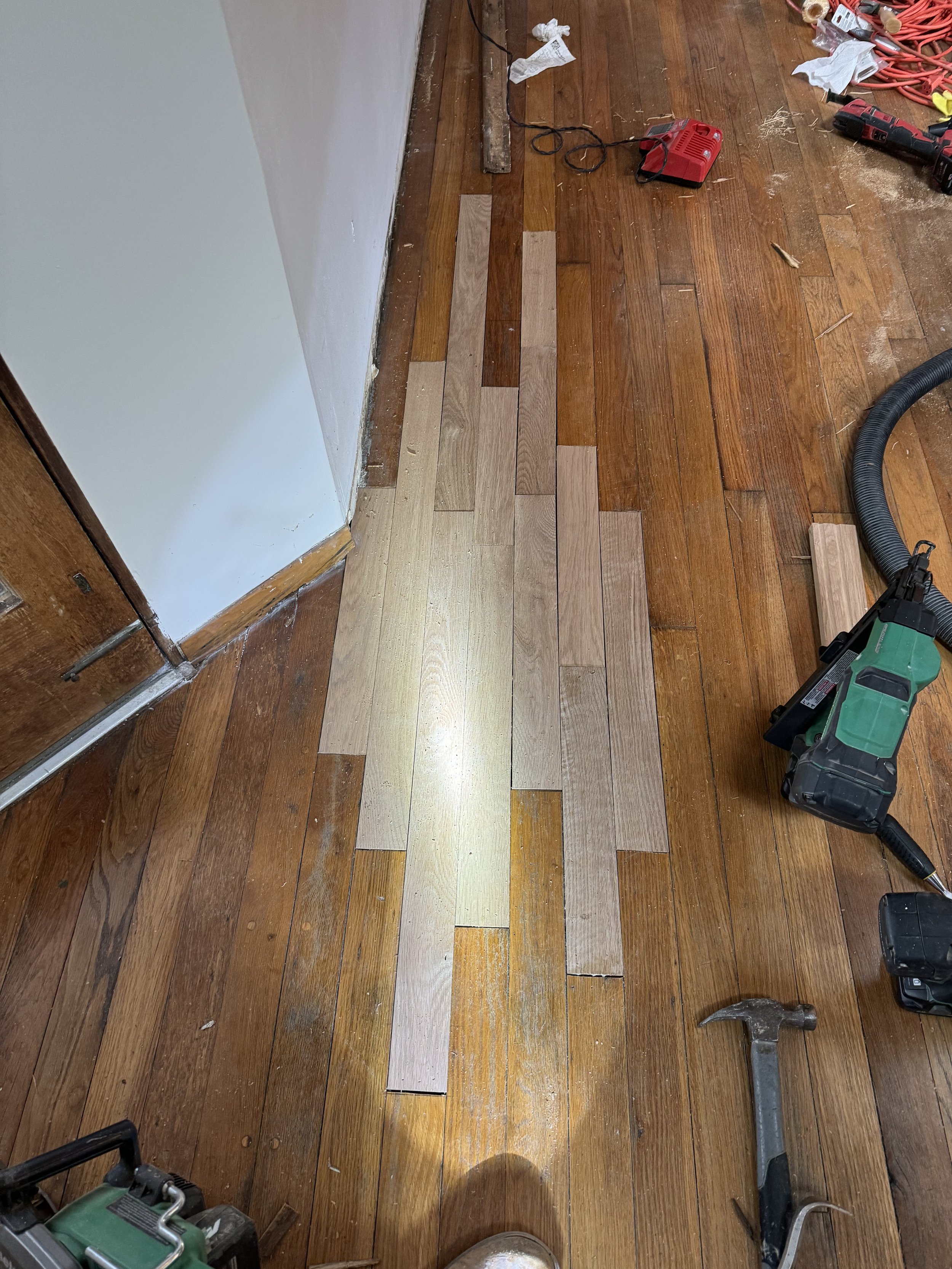 Hardwood floor installation in progress with wooden planks laid out on the floor, various tools including a hammer, drill, and vacuum hose scattered around, and some electrical cords visible.
