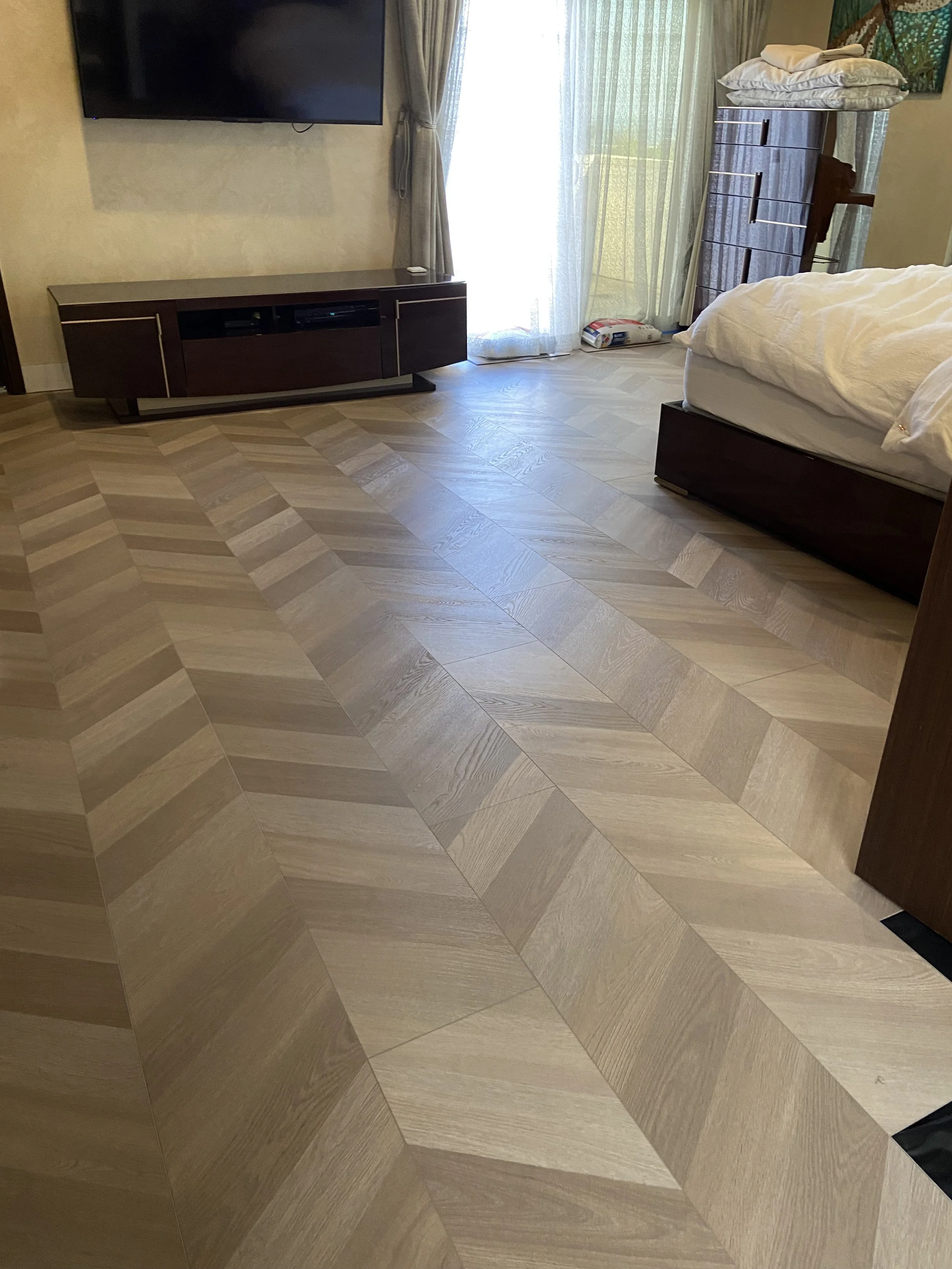 A bedroom with light-colored wooden flooring, a dresser with folded clothes on top, a bed with white bedding, a flat-screen TV mounted on the wall, and a sliding glass door with curtains leading to a balcony.