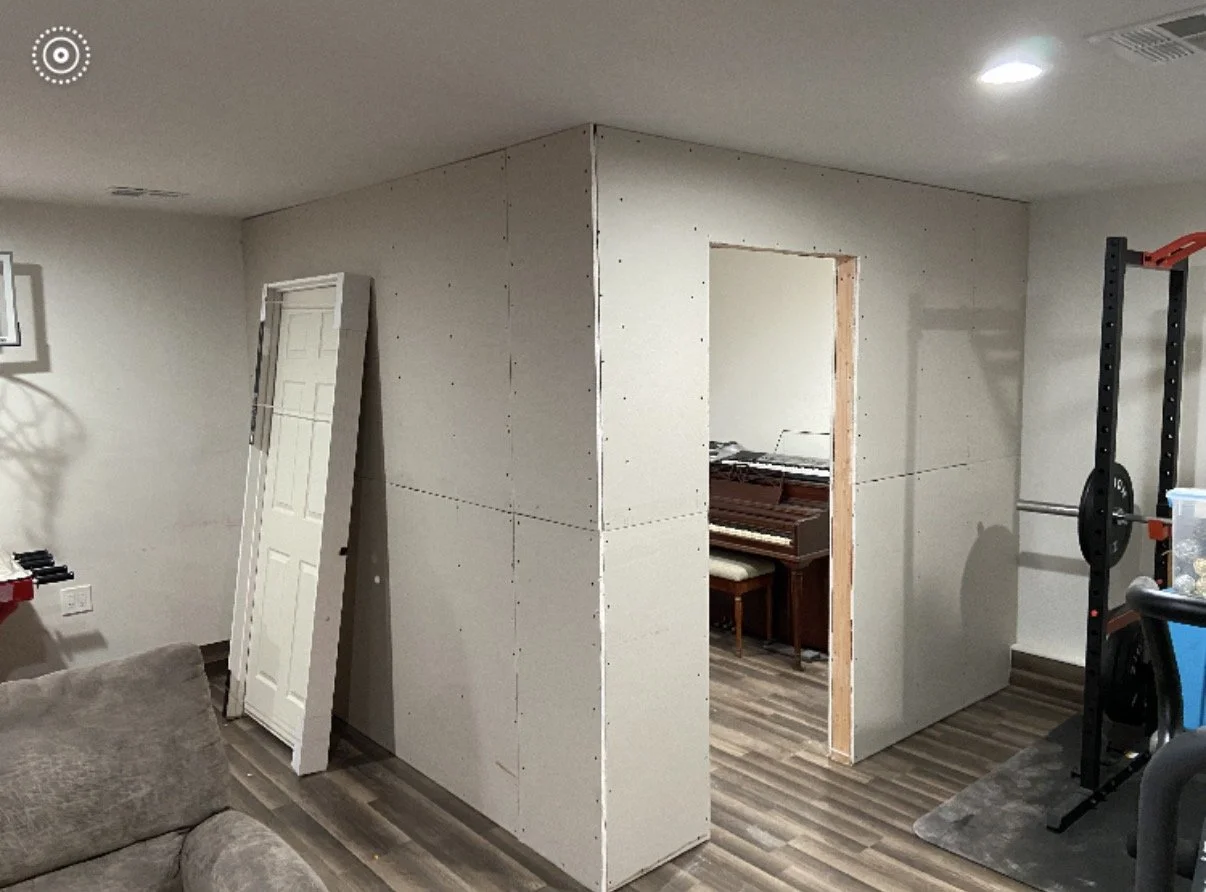 Indoor basement with drywall under construction, a door leaning against the wall, and a small opening in the drywall revealing a room with a piano. There is gym equipment on one side and part of a sofa in the corner.