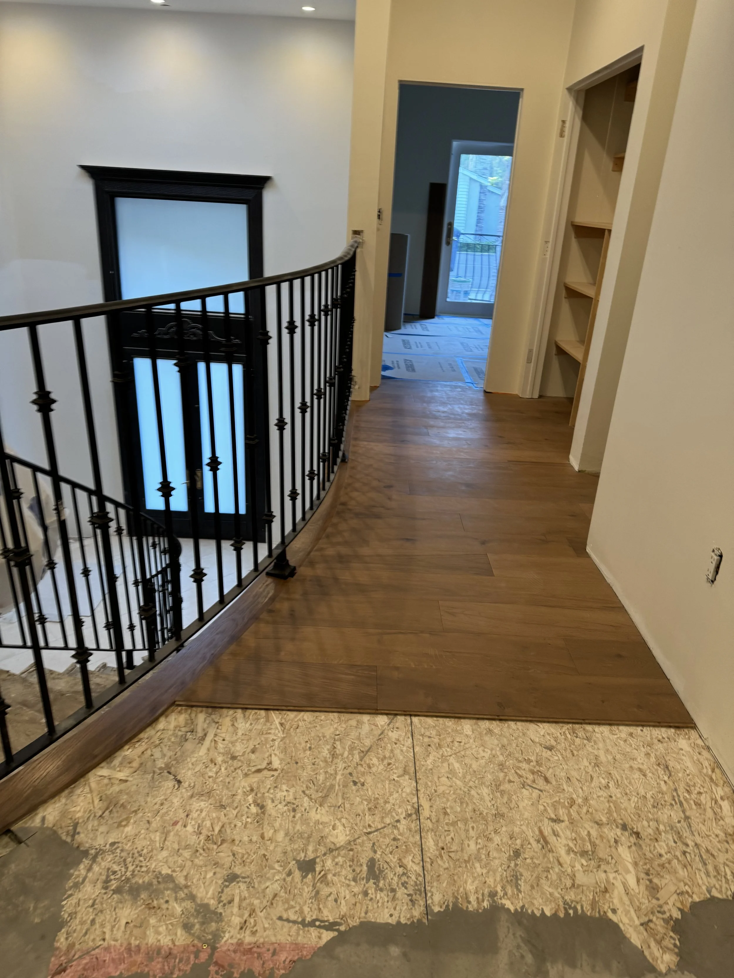 Interior view of a house under construction, showing a staircase with a black metal railing, a wooden floor, a doorway leading to an outdoor balcony, and a partially finished floor with exposed subfloor.