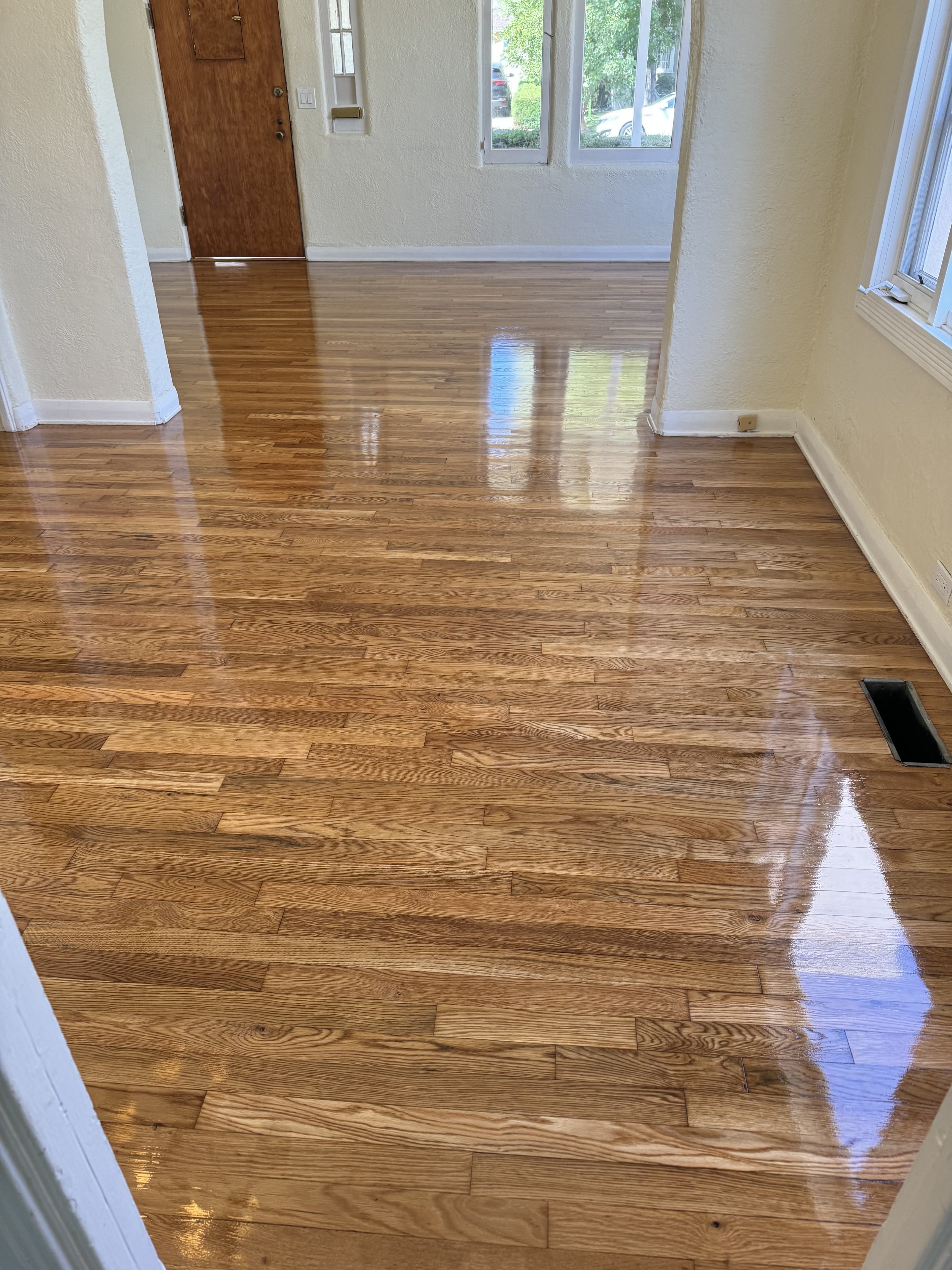 Polished hardwood floors in a bright living room with a doorway, windows, and a view of trees outside.