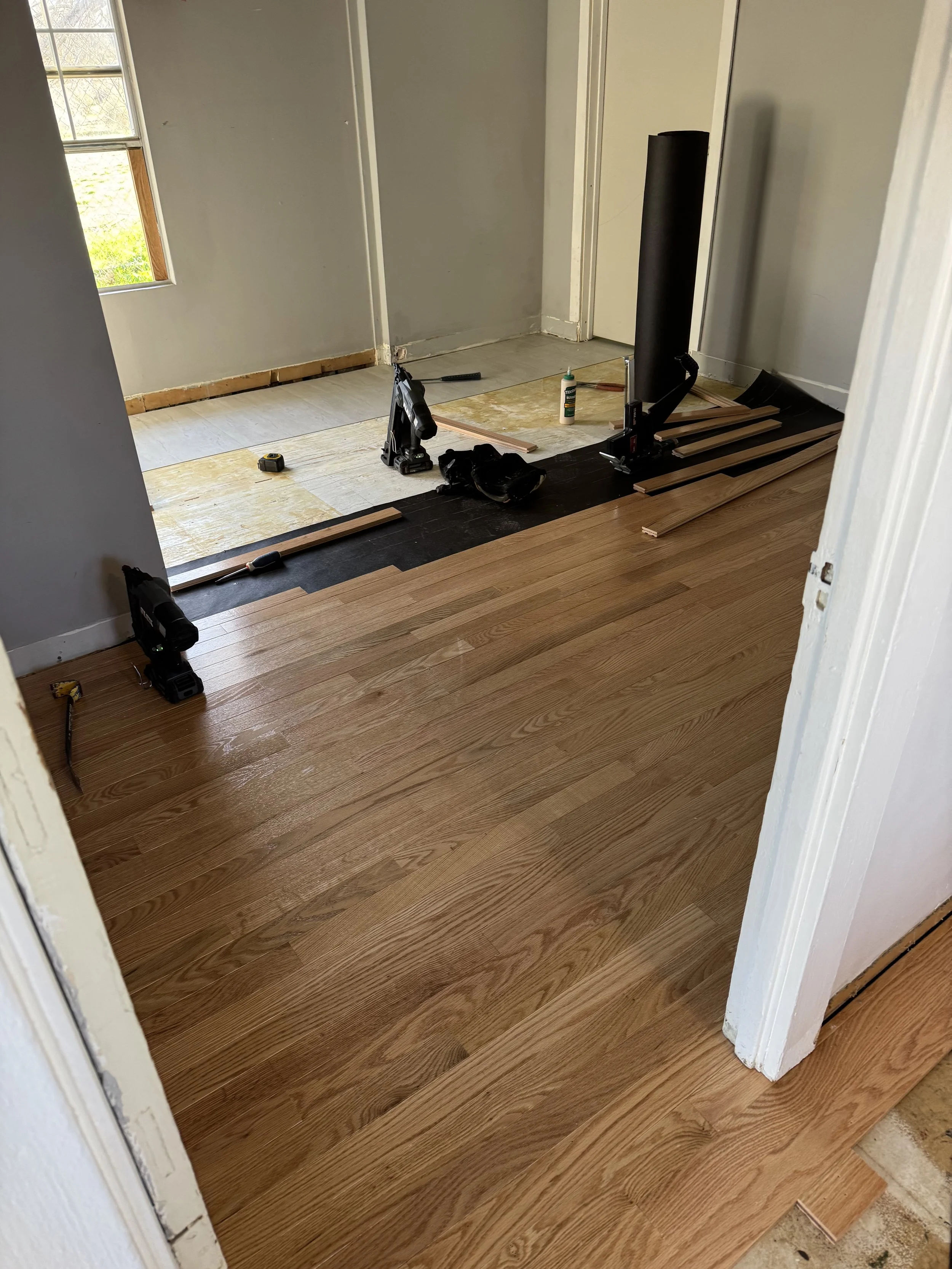 Room with hardwood flooring being installed, tools and supplies like a measuring tape, nail gun, wood planks, adhesive, and a level are visible, with some area still needing flooring.