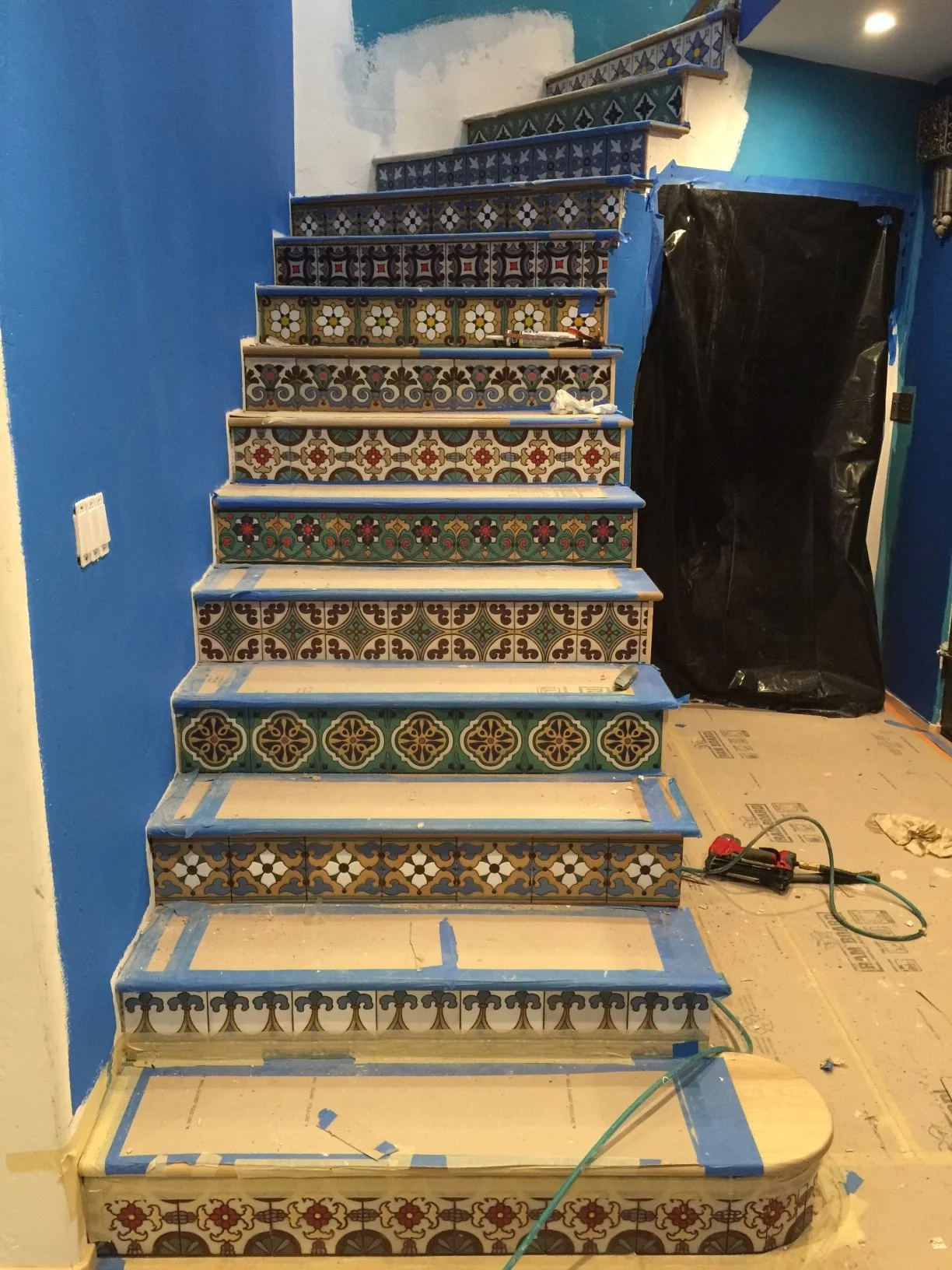 Interior view of a staircase under renovation, featuring decorative colorful tiles on the steps and blue painter's tape along the edges, with visible construction tools and materials around.