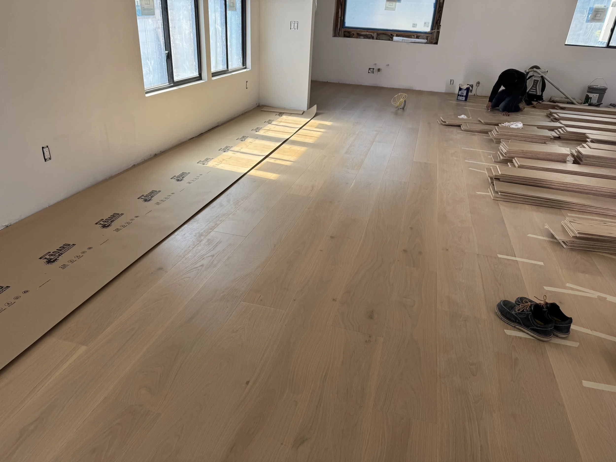 A room under construction with a worker installing wooden flooring, stacks of wooden planks on the right, an open shoe on the floor, and a large window providing natural light.