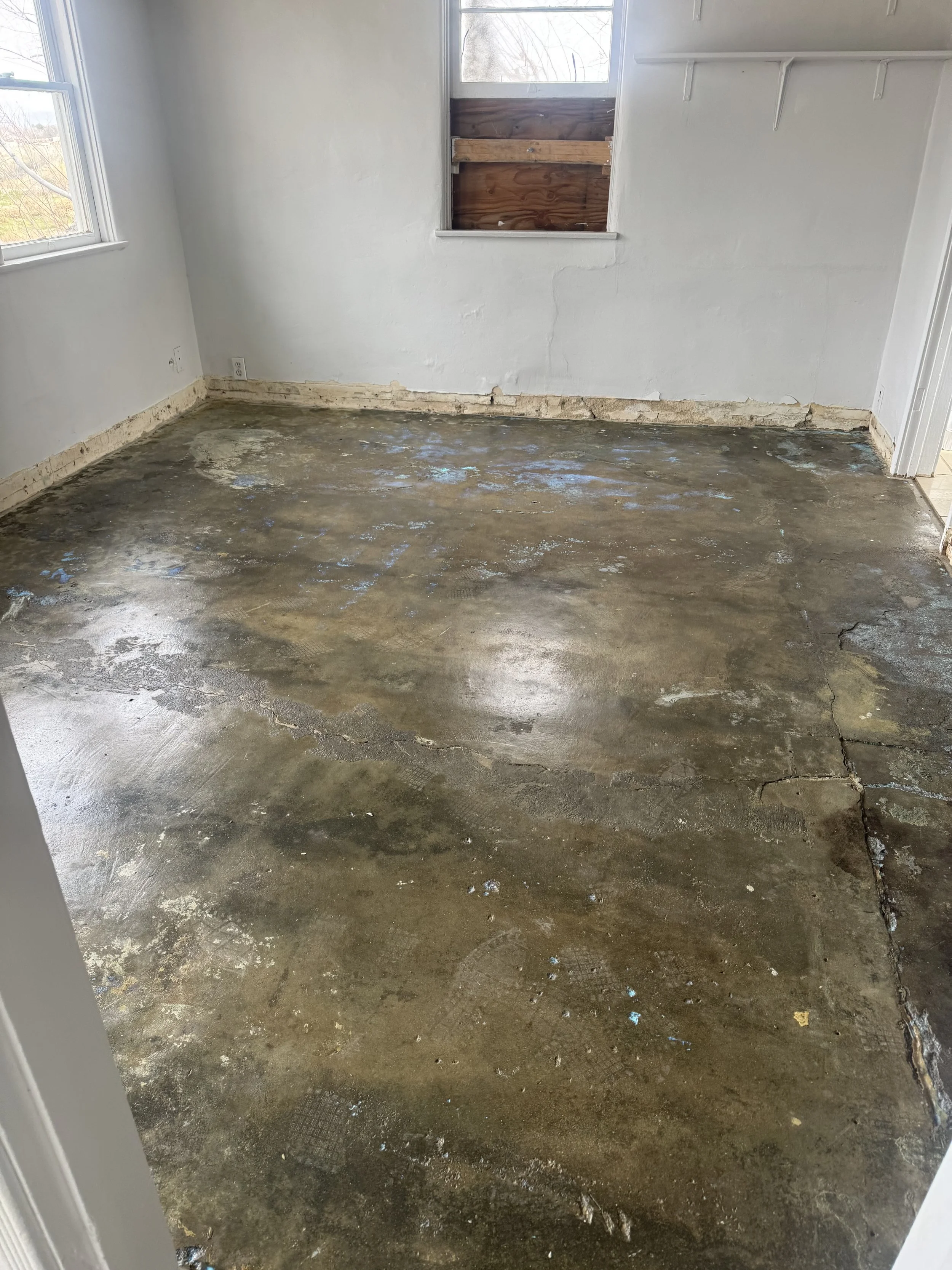 Empty room with concrete floor, white walls, and a window with part of the wall boarded up with wood.