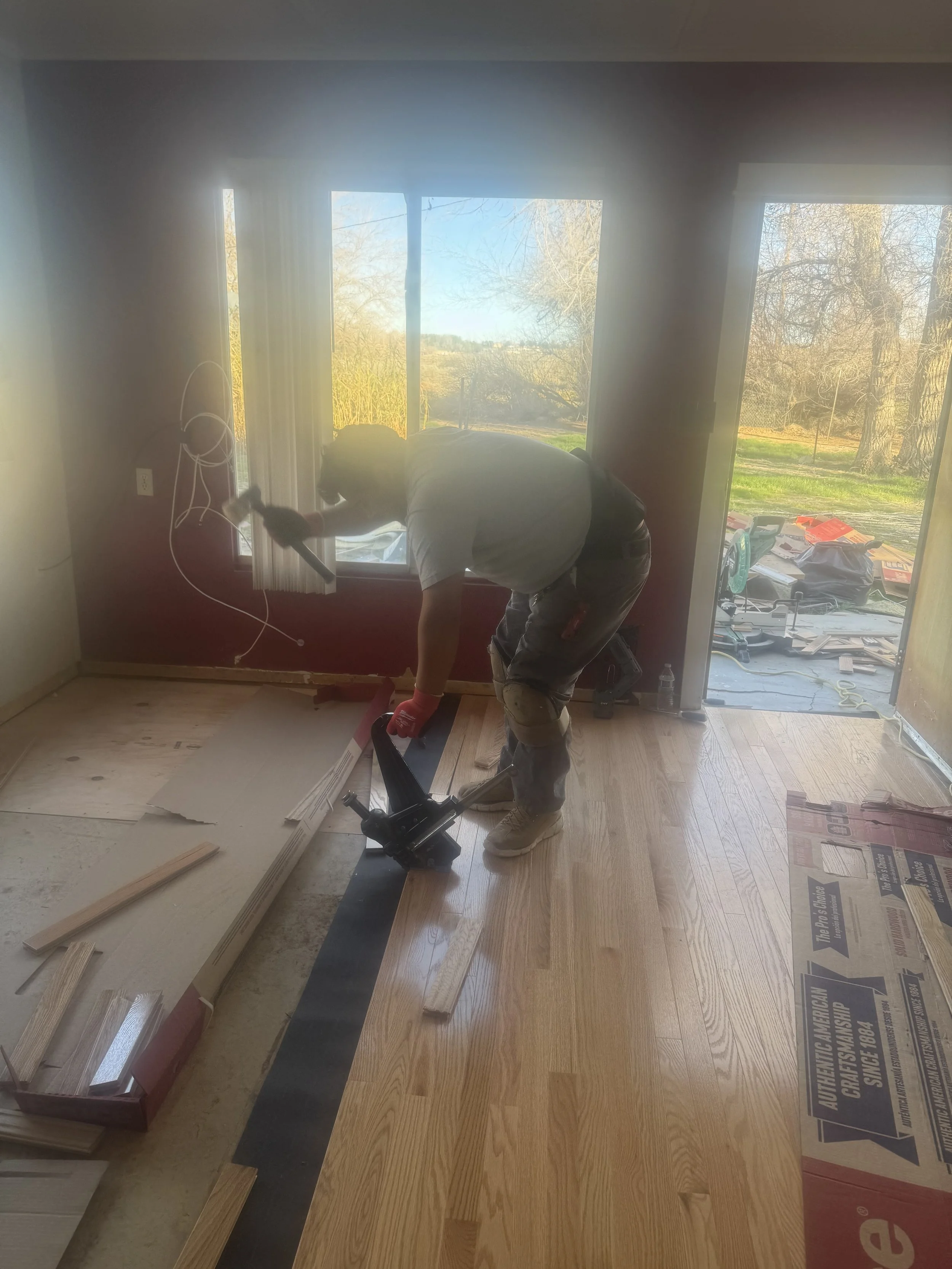 A person installing hardwood flooring in a room with large windows, using a power tool to lay down wood planks.