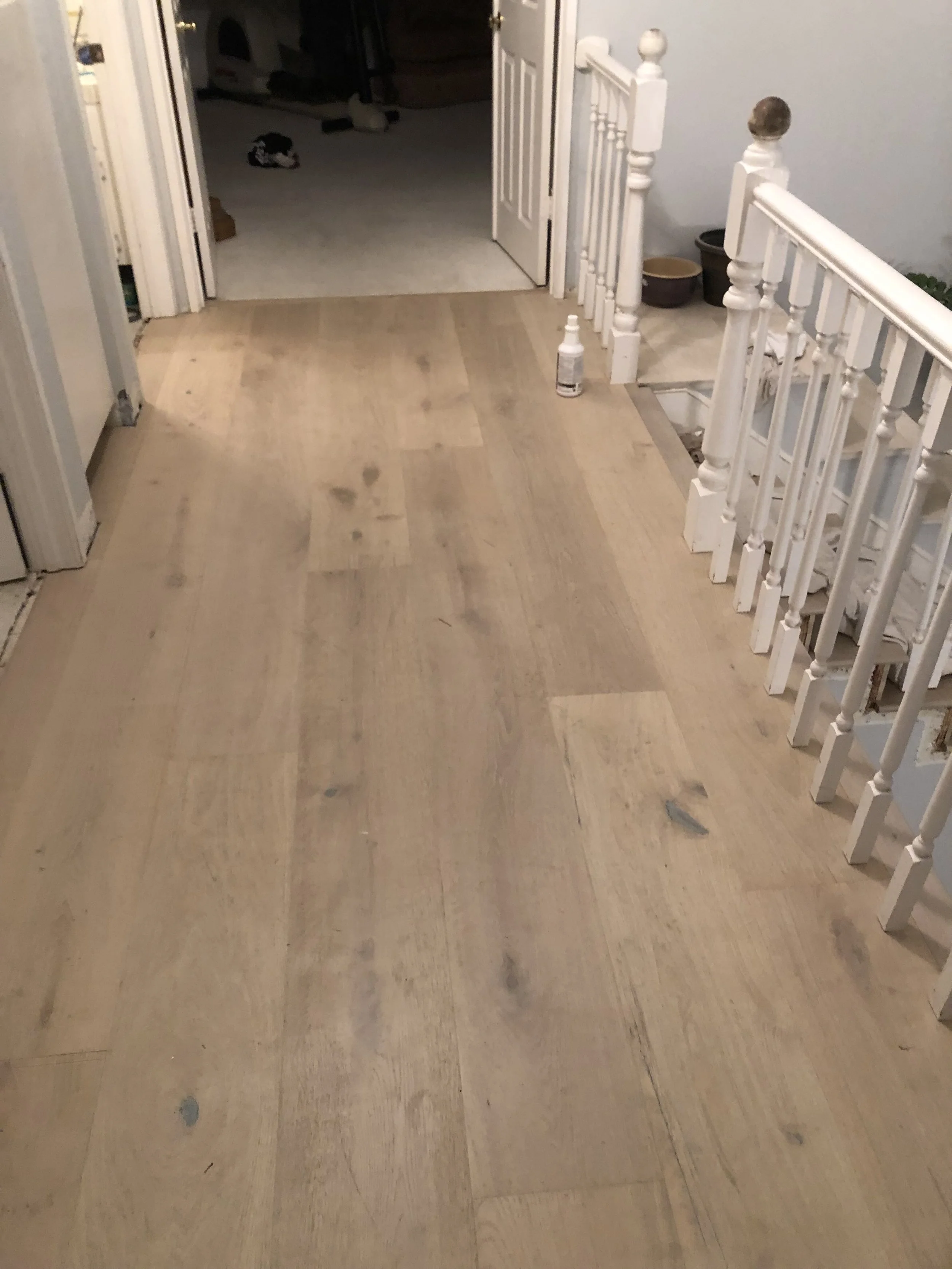 Interior view of a hallway with light-colored hardwood flooring under renovation, with a partially finished section near the stairs and an open door leading to a room with carpeted flooring. Some household items are visible in the background.