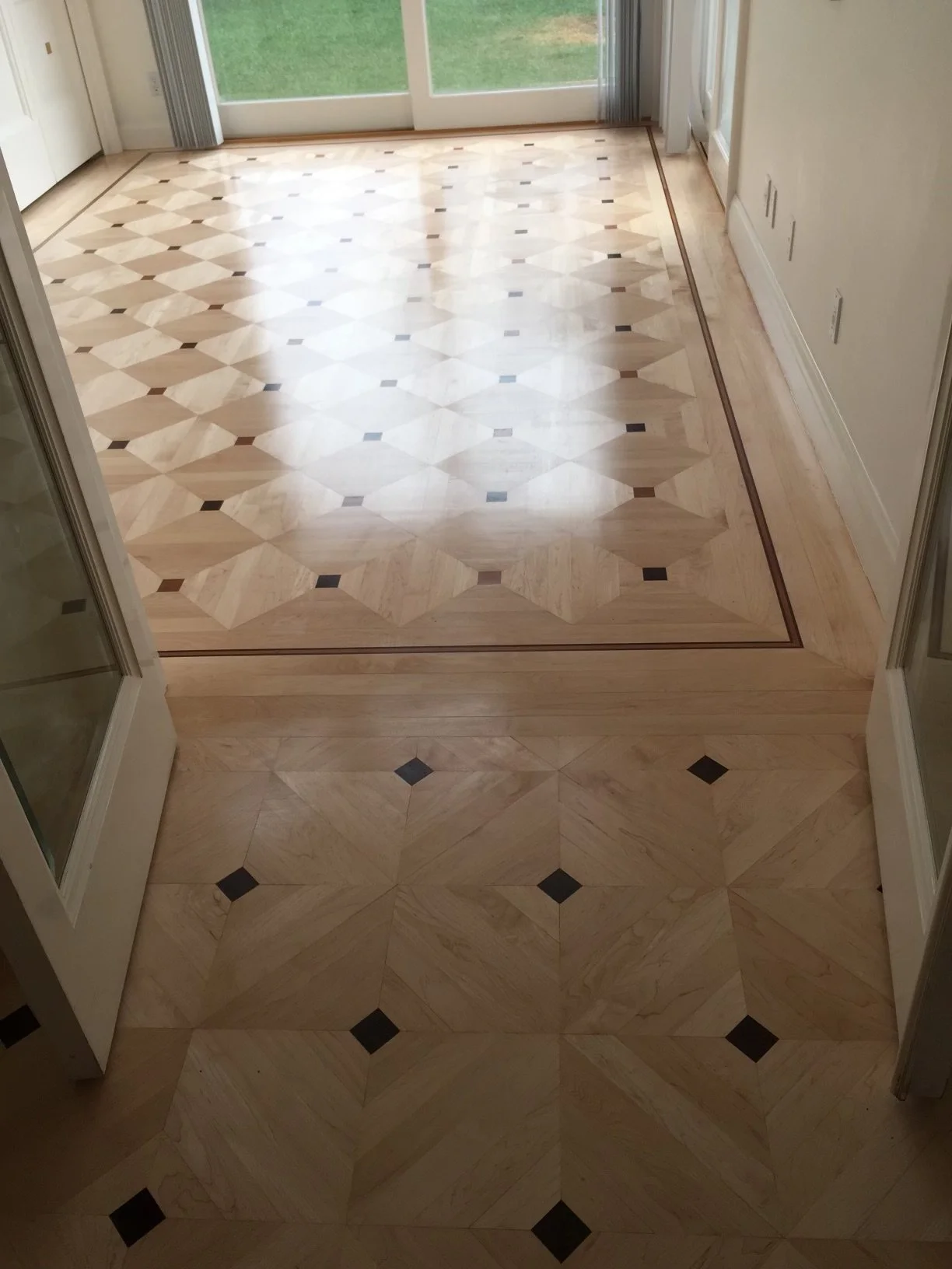 Photo of a room with official-style parquet flooring pattern, featuring beige and black tiles, leading to a glass sliding door with a view of a backyard.