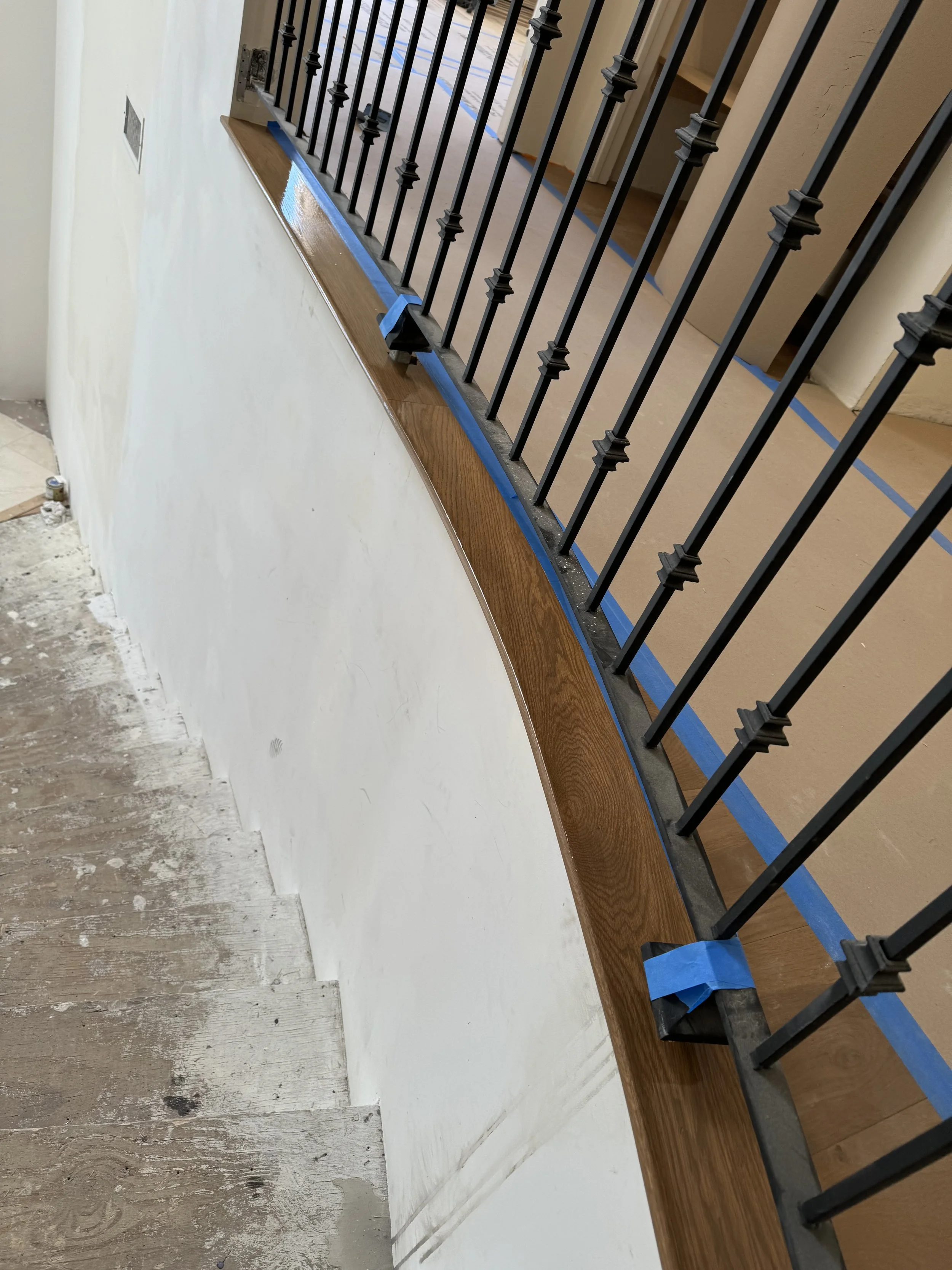 A staircase railing with black metal balusters, a wooden handrail, and a wall beneath, with painter's tape protecting the edges during renovation.