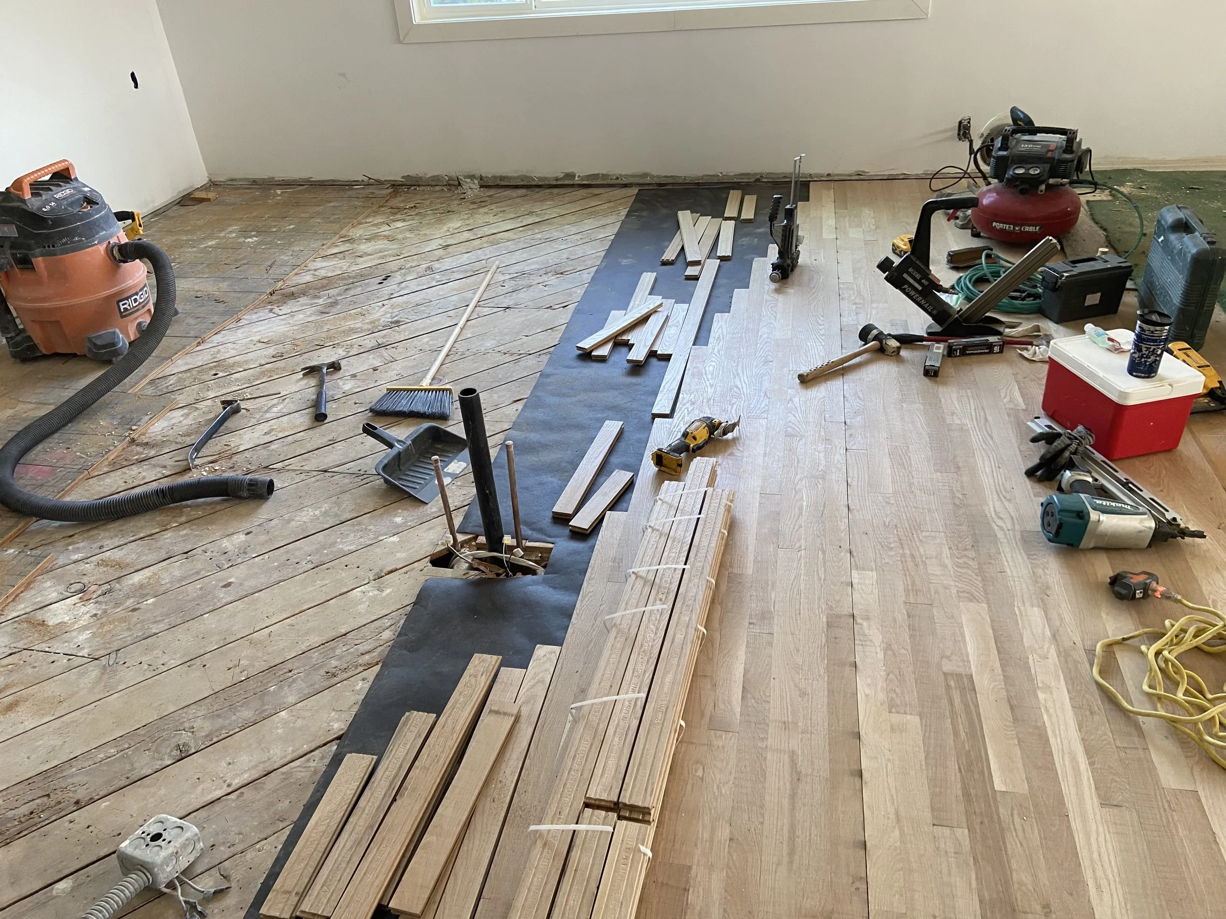 Room under renovation with hardwood flooring being installed, tools, wood planks, and construction equipment scattered around.