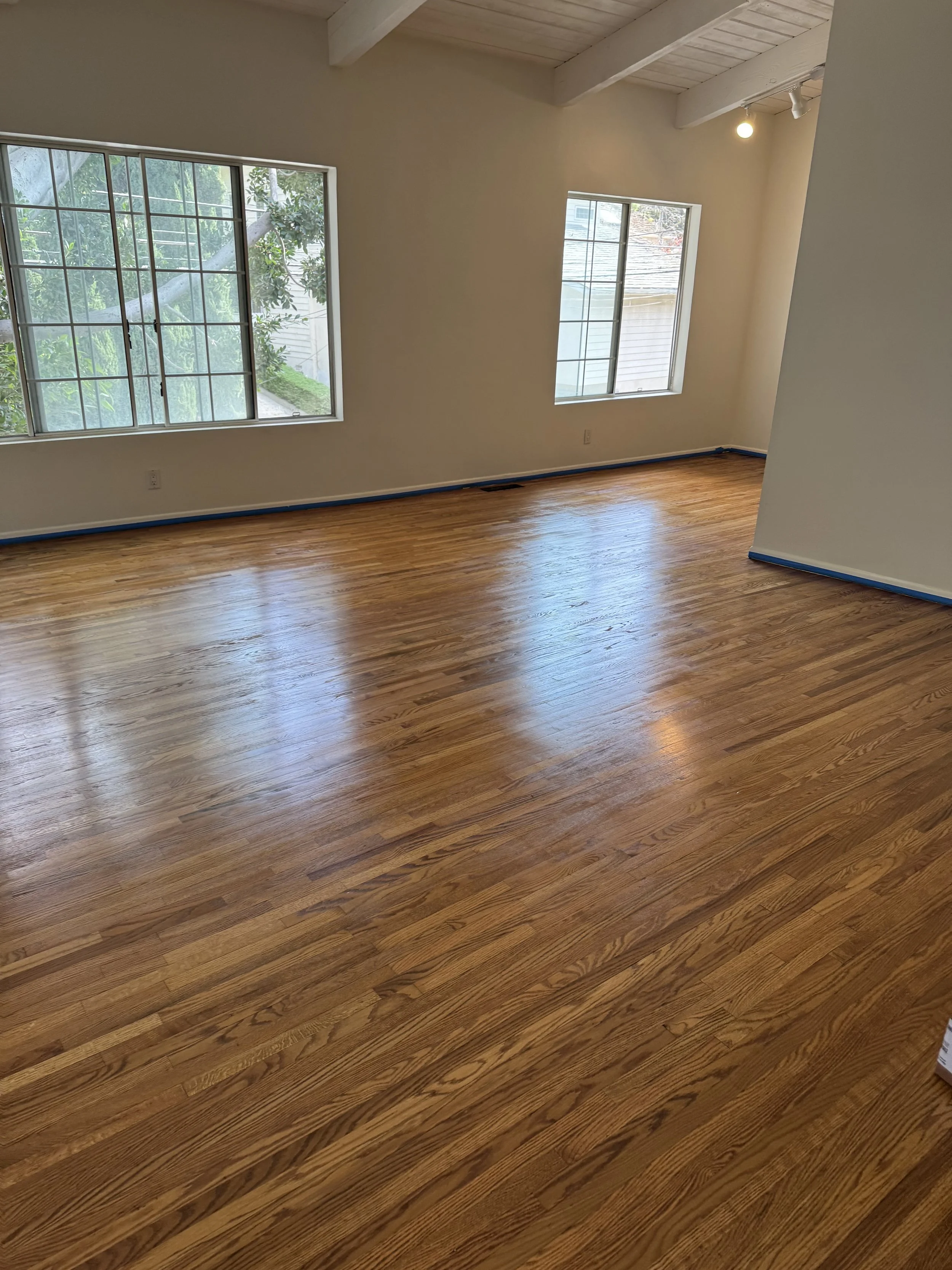 Bright empty room with hardwood floors, white walls, two large windows, and exposed ceiling beams.