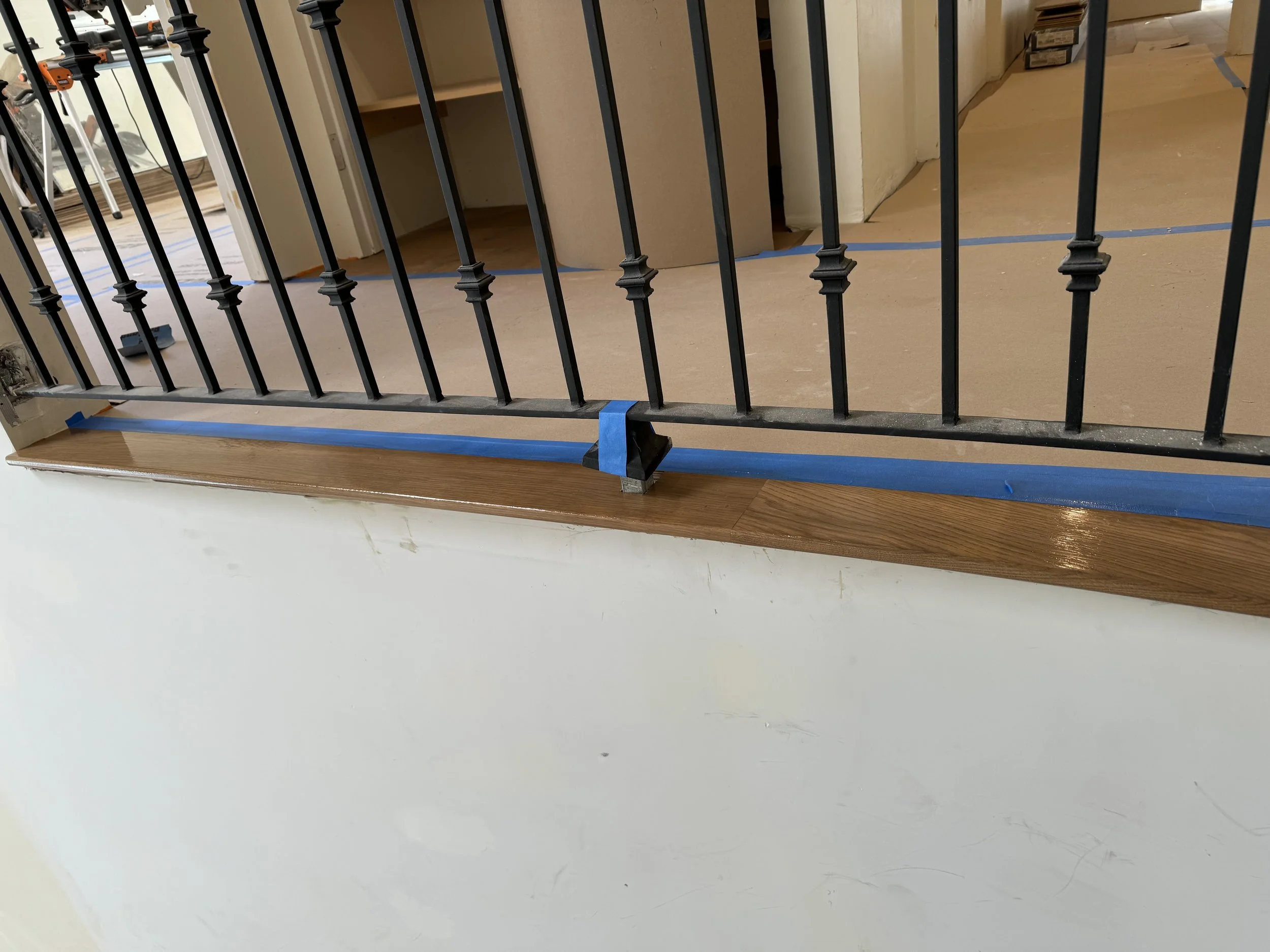 Close-up of a black metal railing with wooden handrail, installed on a white wall, with blue painter's tape temporarily securing the railing during construction.