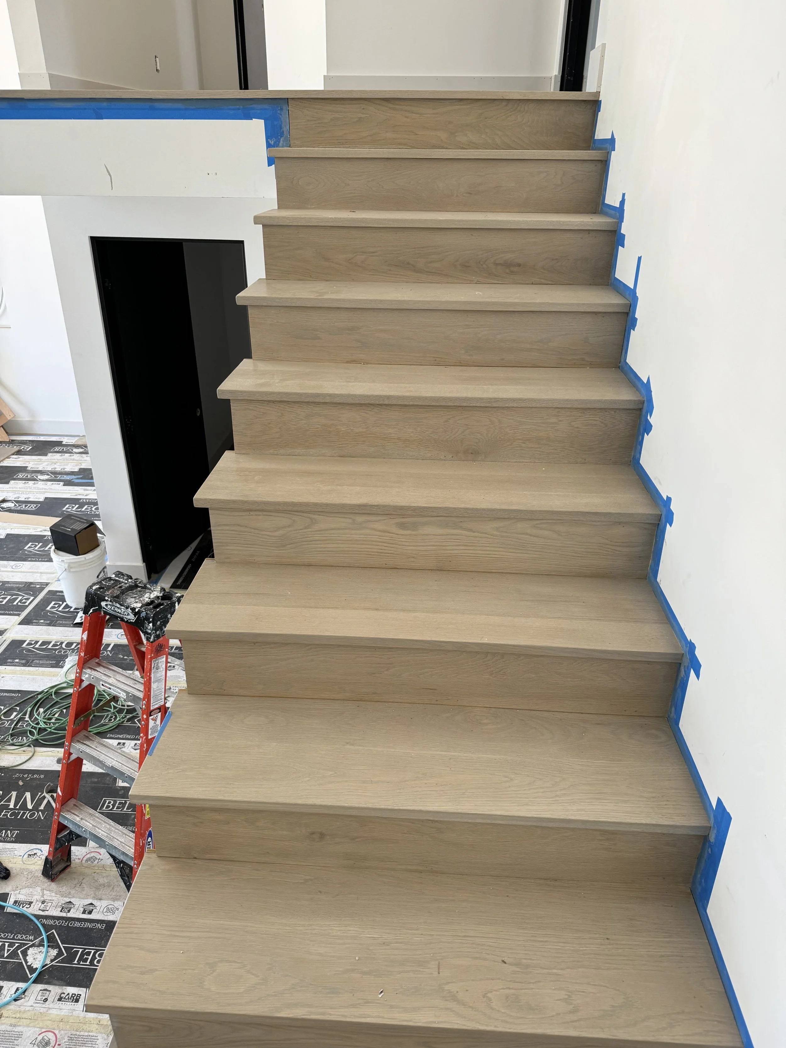 Wooden staircase under construction with blue painter's tape along the edges of the wall, construction tools, ladder, and protective covering on the floor.