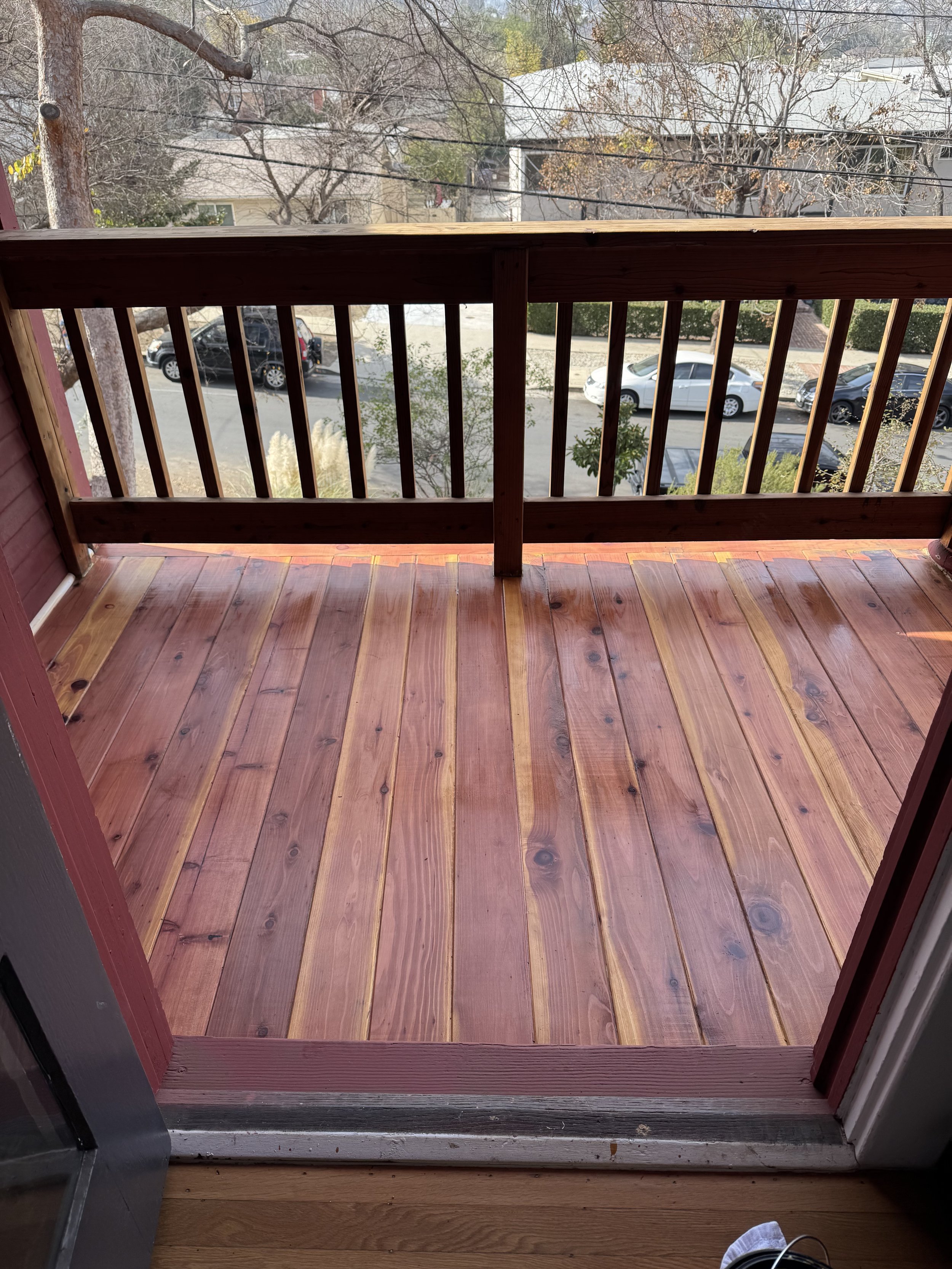 View of a small wooden balcony with a railing overlooking a parking lot with cars and trees.