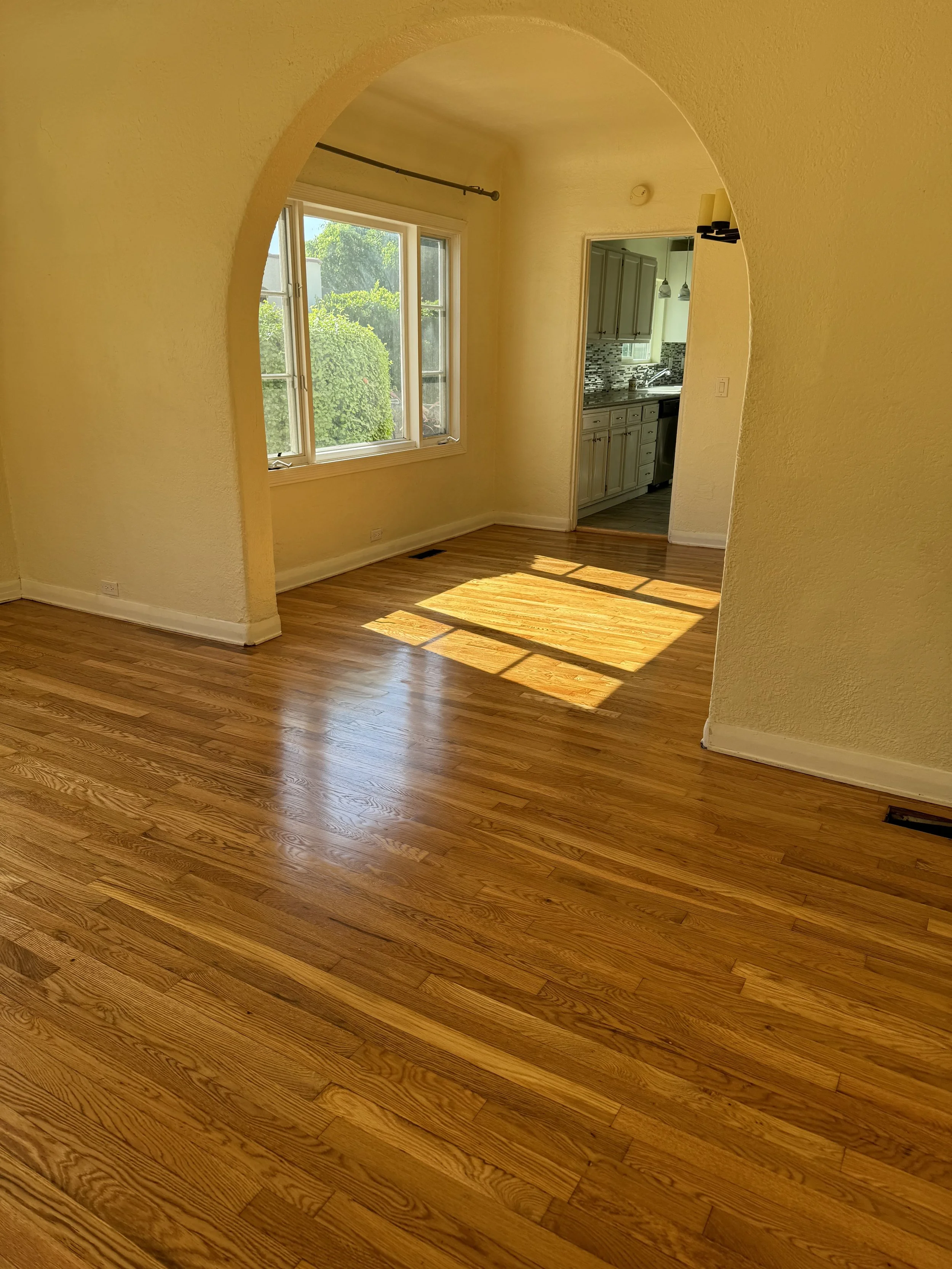 Empty room with hardwood floors, a large window letting in sunlight, and an arched doorway leading to a kitchen with white cabinets and a mosaic tile backsplash.