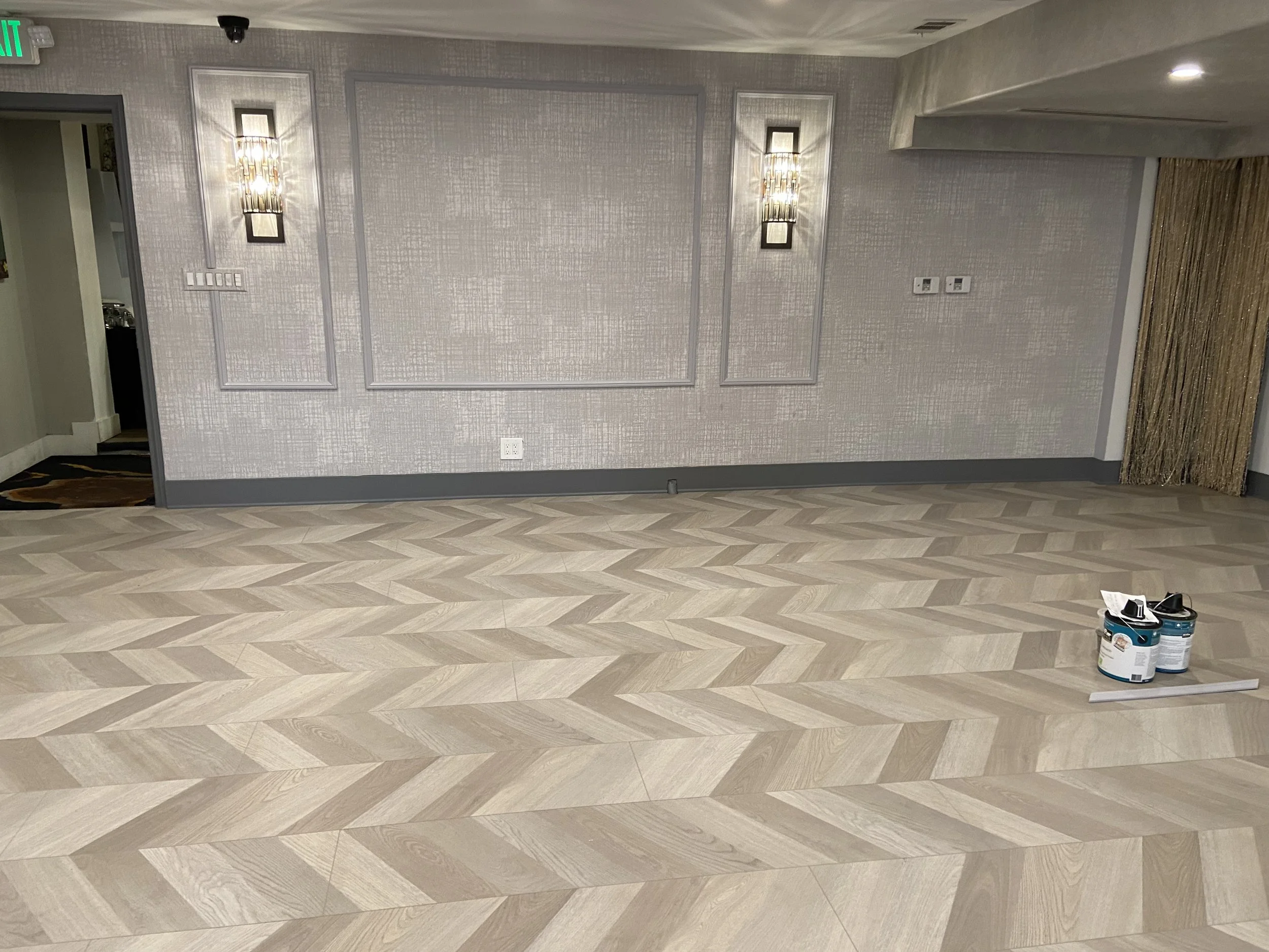 Empty room with patterned light-colored wooden floor, textured wallpapered walls, wall sconces, and construction supplies on the floor.