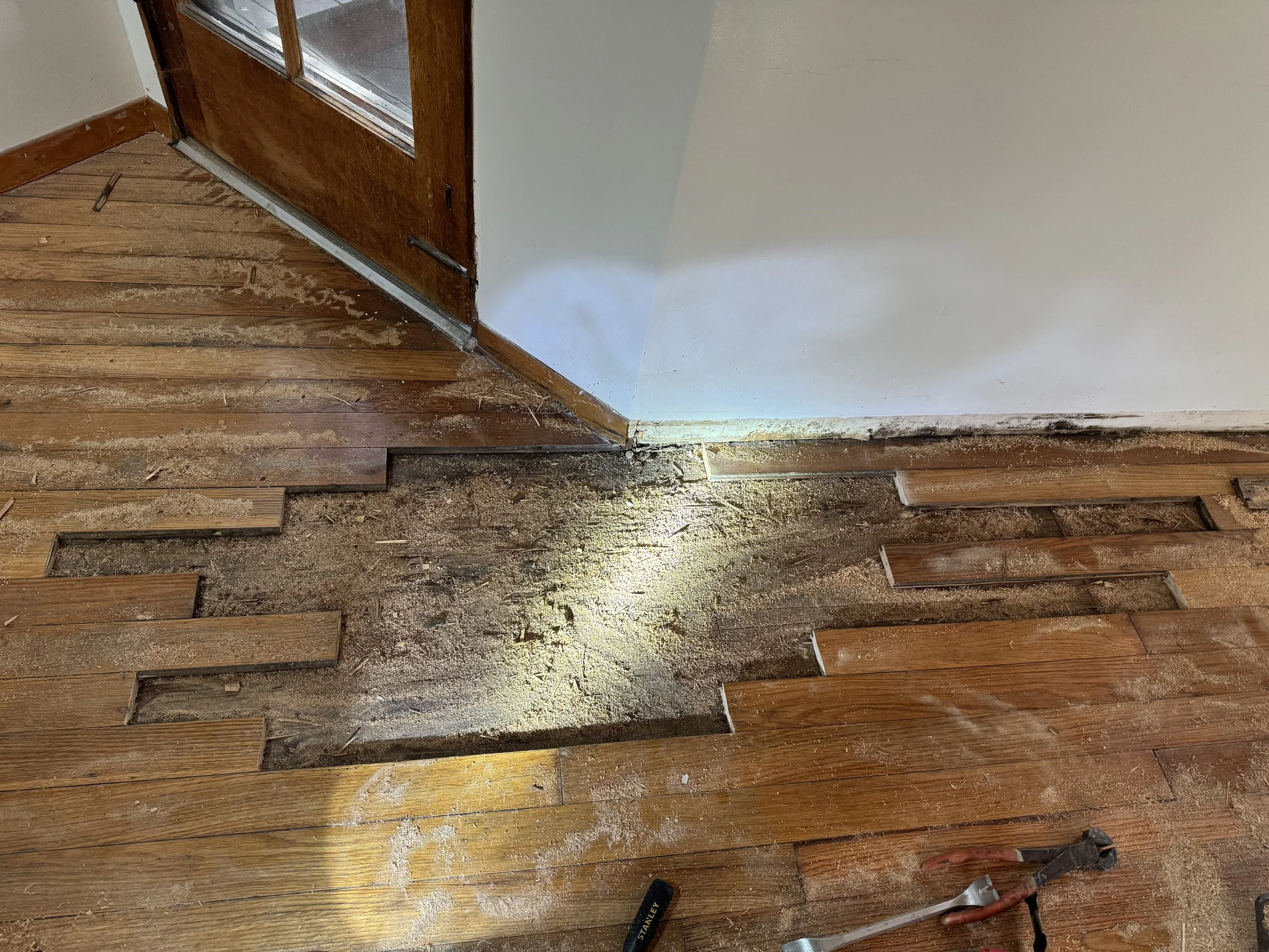 A section of wooden floor with some tiles removed, exposing subfloor and dirt underneath. Tools are on the floor near the area.