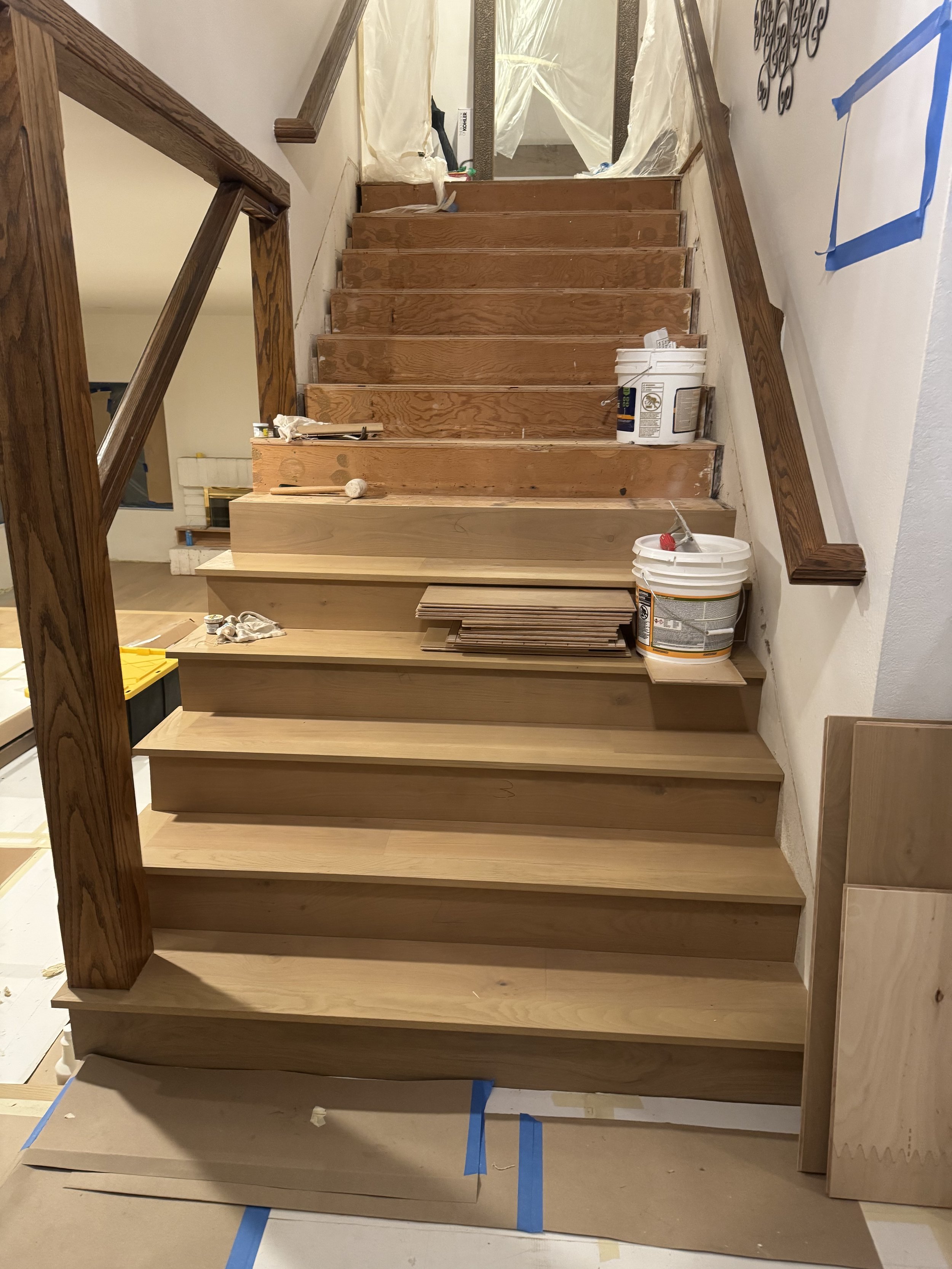 Interior view of a staircase under construction, with newly installed wooden steps and ongoing work materials such as paint buckets, tiles, and tools.