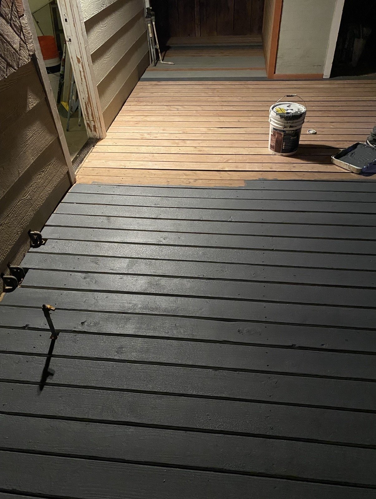 Partially painted wooden deck with gray paint in the foreground and natural wood color in the background, a bucket of paint, and painting supplies on the deck, and a door opening into a room.