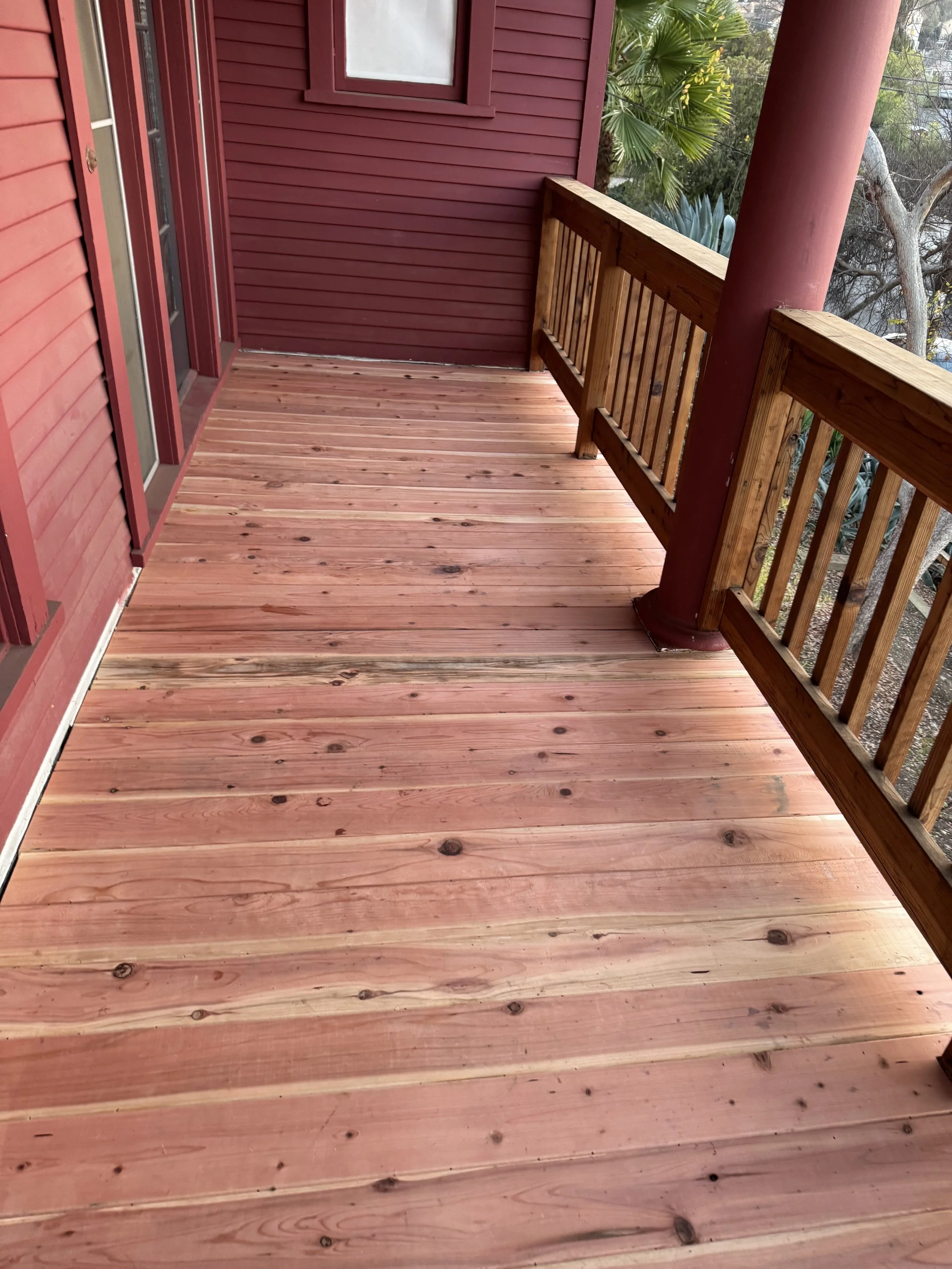 Freshly built wooden balcony with pinkish wood planks, red painted siding, and matching red pillars, overlooking trees.