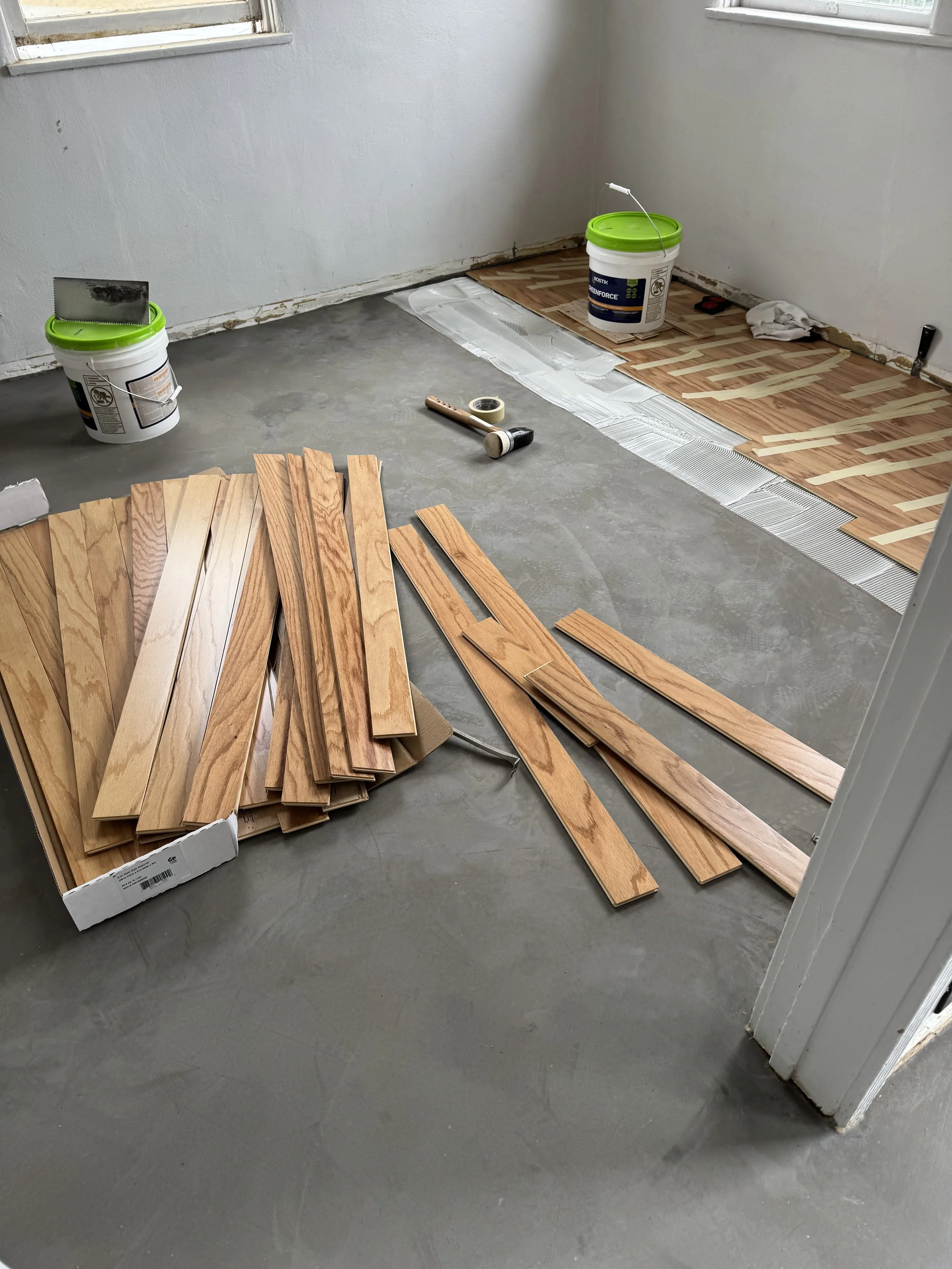 Room under renovation with hardwood flooring being installed, wooden planks, buckets of adhesive, a mallet, tape, and tools scattered around.