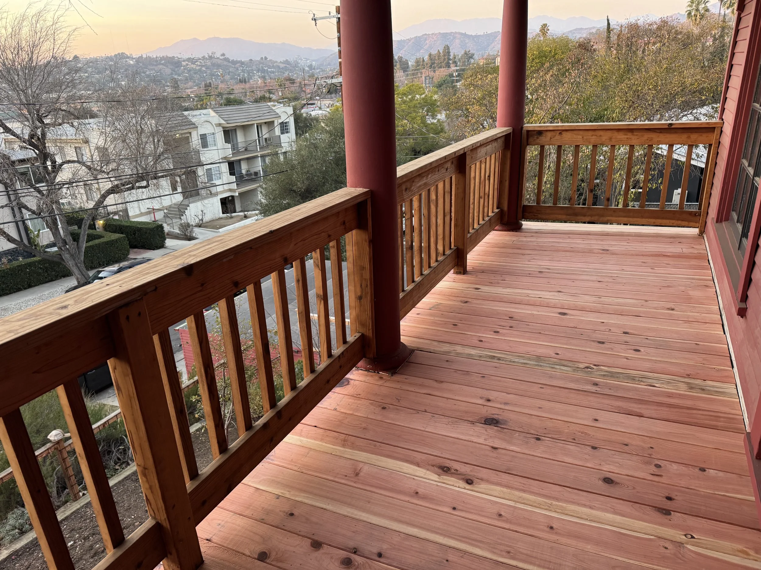 Empty wooden balcony with red railings and posts overlooking residential area and mountains in the distance during sunset.
