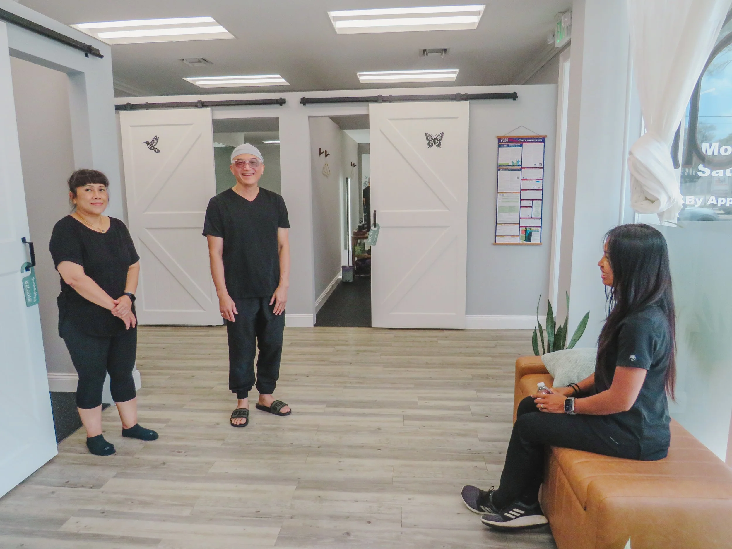 Four people in a waiting area, with two standing and two sitting, inside a modern office or clinic. White sliding doors with butterfly decorations are behind two of them. The space has wood flooring, bright lighting from ceiling fixtures, and a window with sheer curtains.