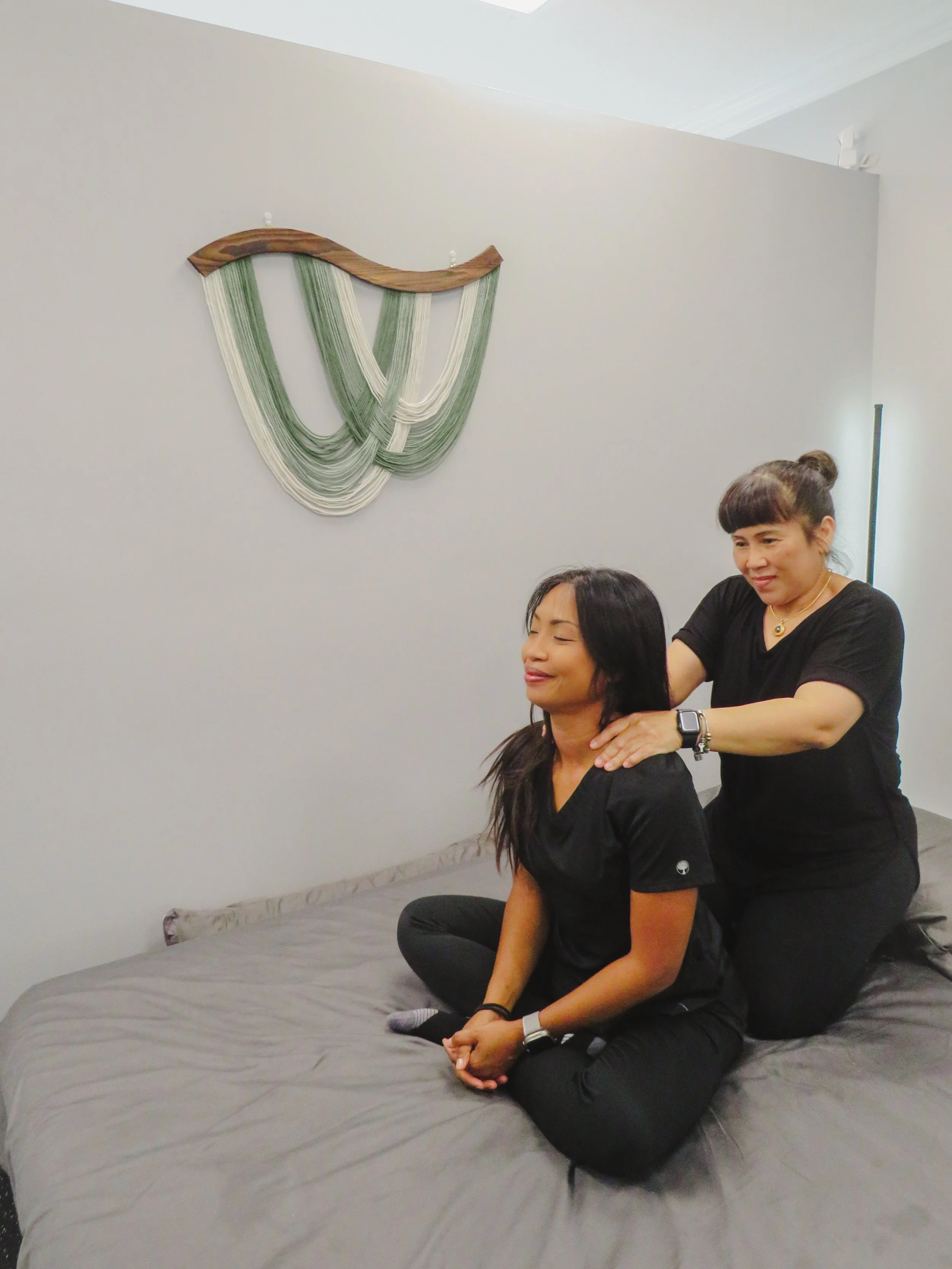 A woman is receiving a seated massage from another woman in a room with a large wall hanging and a bed.