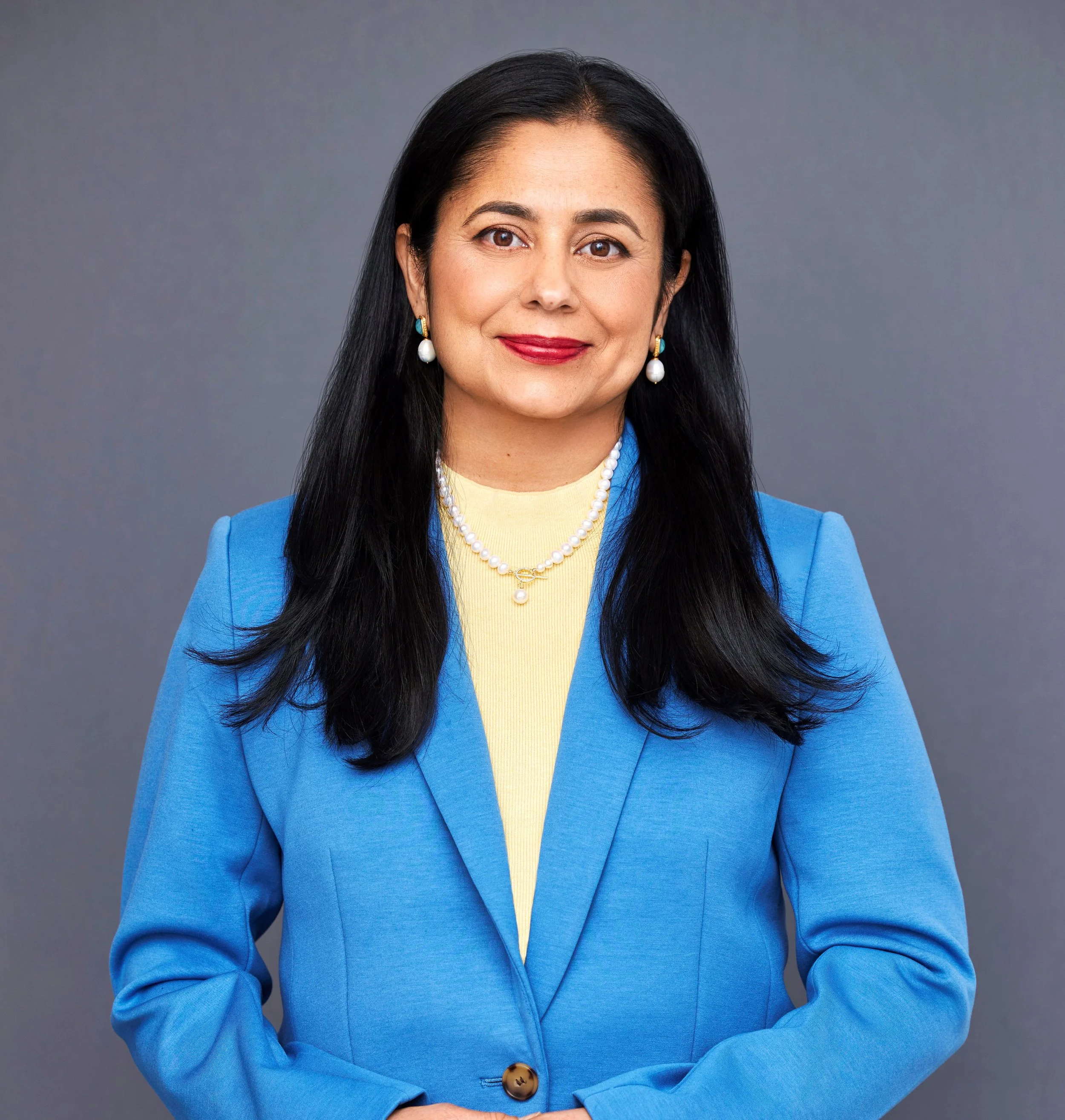 A woman with long black hair, wearing a blue blazer, yellow top, pearl jewelry, and red lipstick, standing against a gray background.