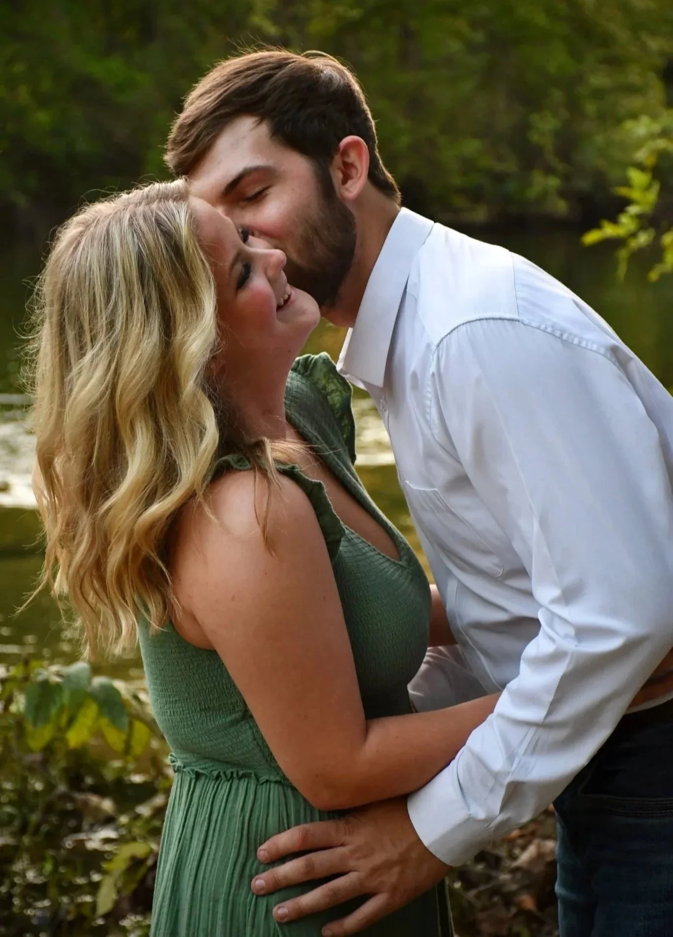 A man and woman close together outdoors near a body of water, smiling. The man is leaning in and kissing the woman's forehead while she touches his arm. They are in nature with trees in the background.
