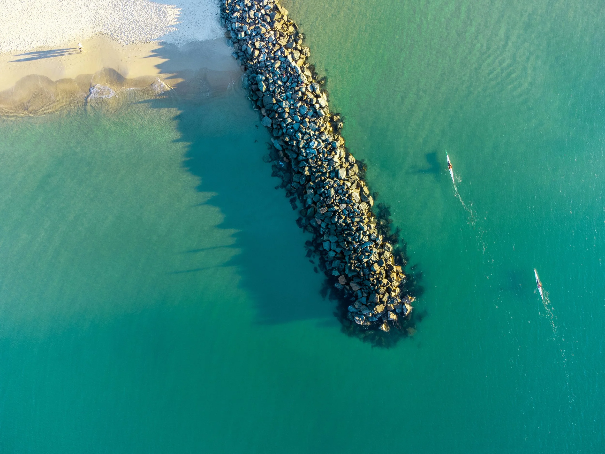 Aerial view of a breakwater made of large rocks extending into the ocean, with a sandy beach on the left and two kayakers on the water to the right.