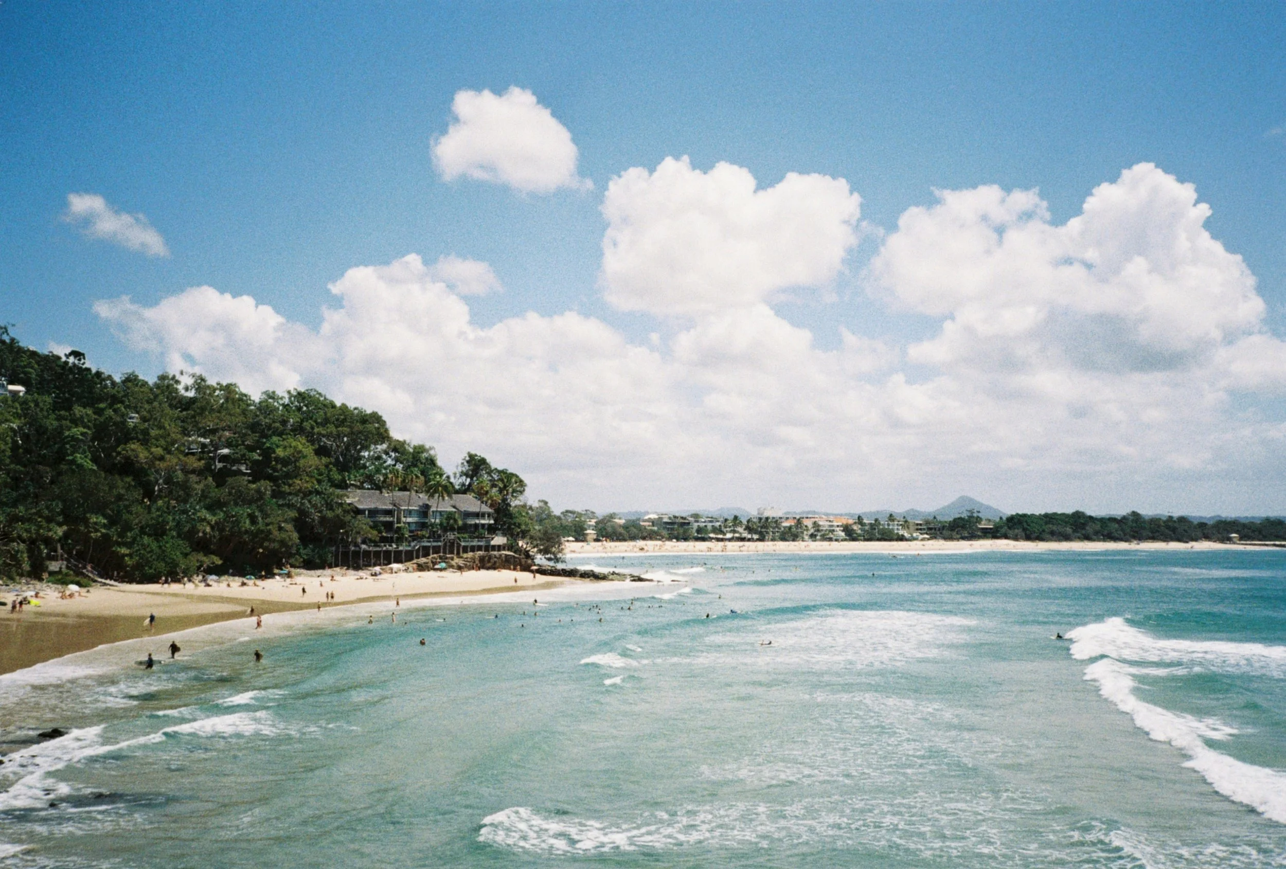 A beach with clear blue water, people swimming and relaxing on the sandy shore, green trees and houses along the coastline, and a partly cloudy sky with fluffy white clouds.