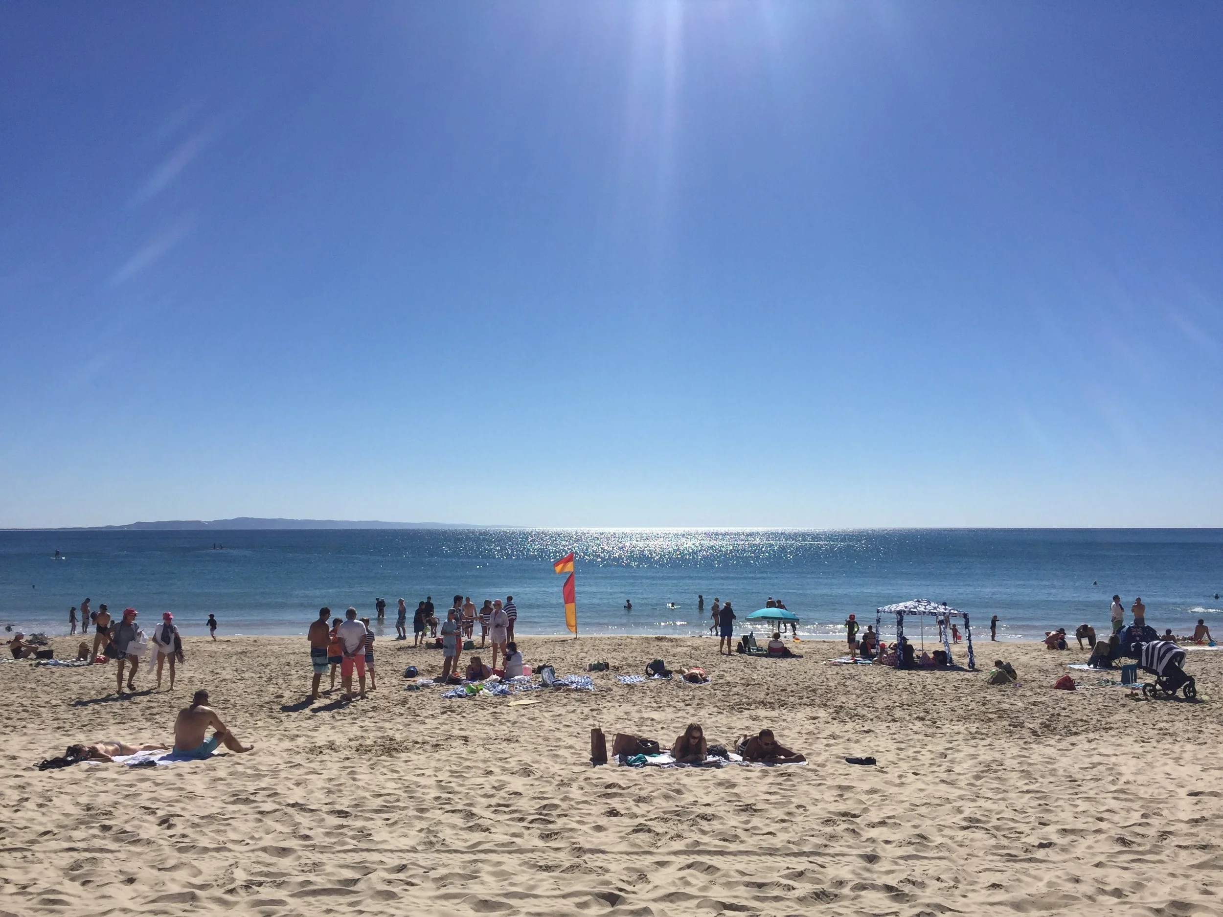 A sunny beach scene with people relaxing on the sand, some under umbrellas, and others swimming or standing in the water. The ocean is calm, with a bright sun overhead, and distant land on the horizon.