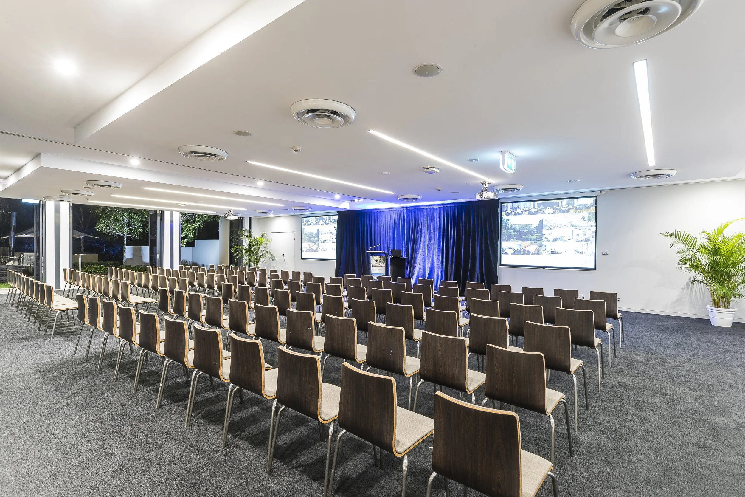 Empty conference room set up for a presentation, with rows of chairs facing a stage with a podium, curtains, and large screens on the wall.