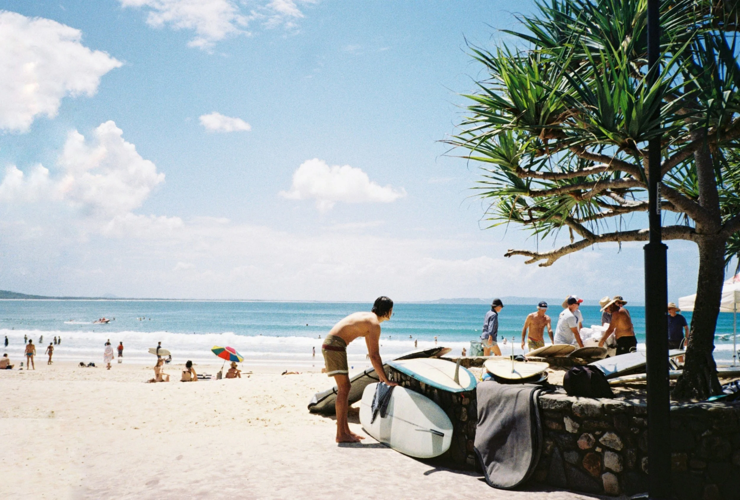 Beach scene with people relaxing, beach equipment, and surfboards near a tree under a partly cloudy sky.
