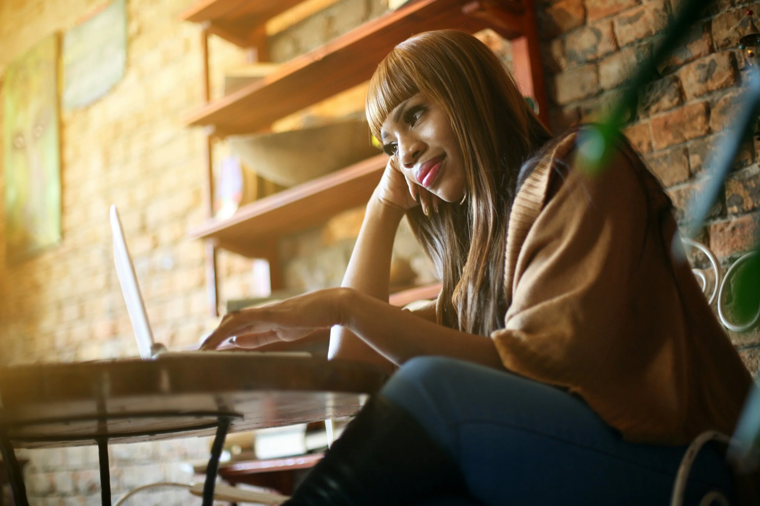 A woman with long, straight hair and dark makeup sitting at a wooden table, smiling while looking at her phone in a cozy cafe with brick walls and wooden shelves.