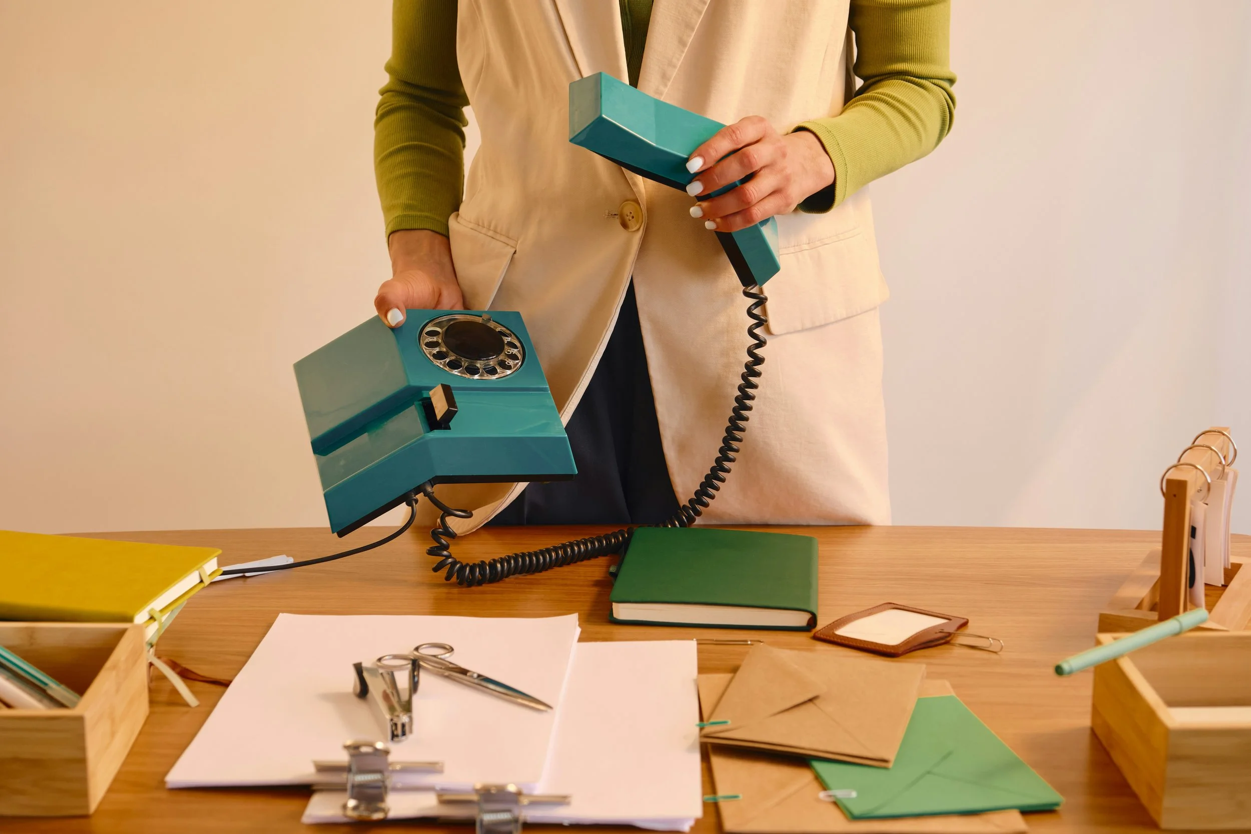 A person dressed in a beige vest and green long-sleeve shirt holding a turquoise vintage rotary phone with a cord, standing behind a wooden desk with various office supplies, papers, notebooks, scissors, and a stapler scattered across the surface.