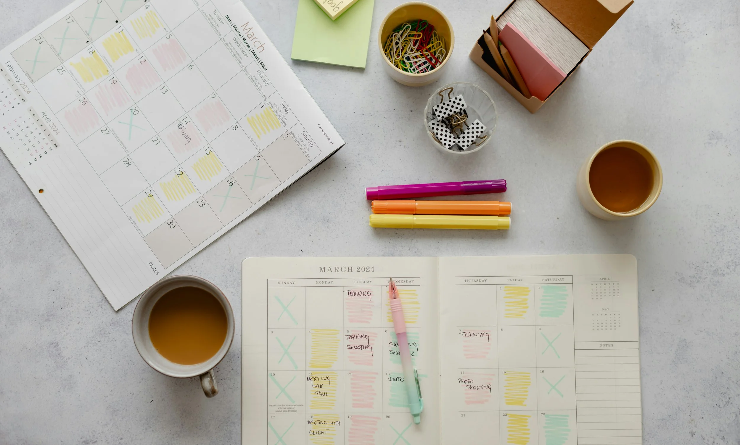 A desk with March and April 2024 calendars, notes, pens, sticky notes, a box of colored paperclips, a small container with black and white dice, two cups of coffee, and a cup of paper clips, arranged on a white surface.