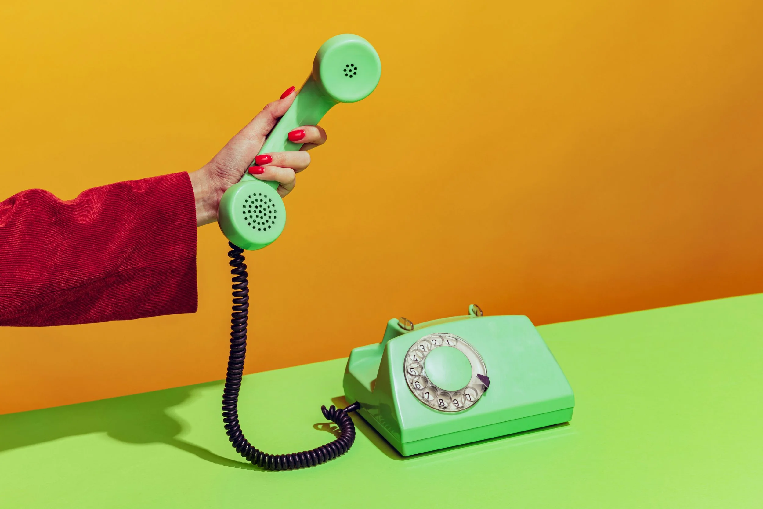 A person holding a green vintage rotary telephone receiver above a matching green landline base on a green table, with an orange background.