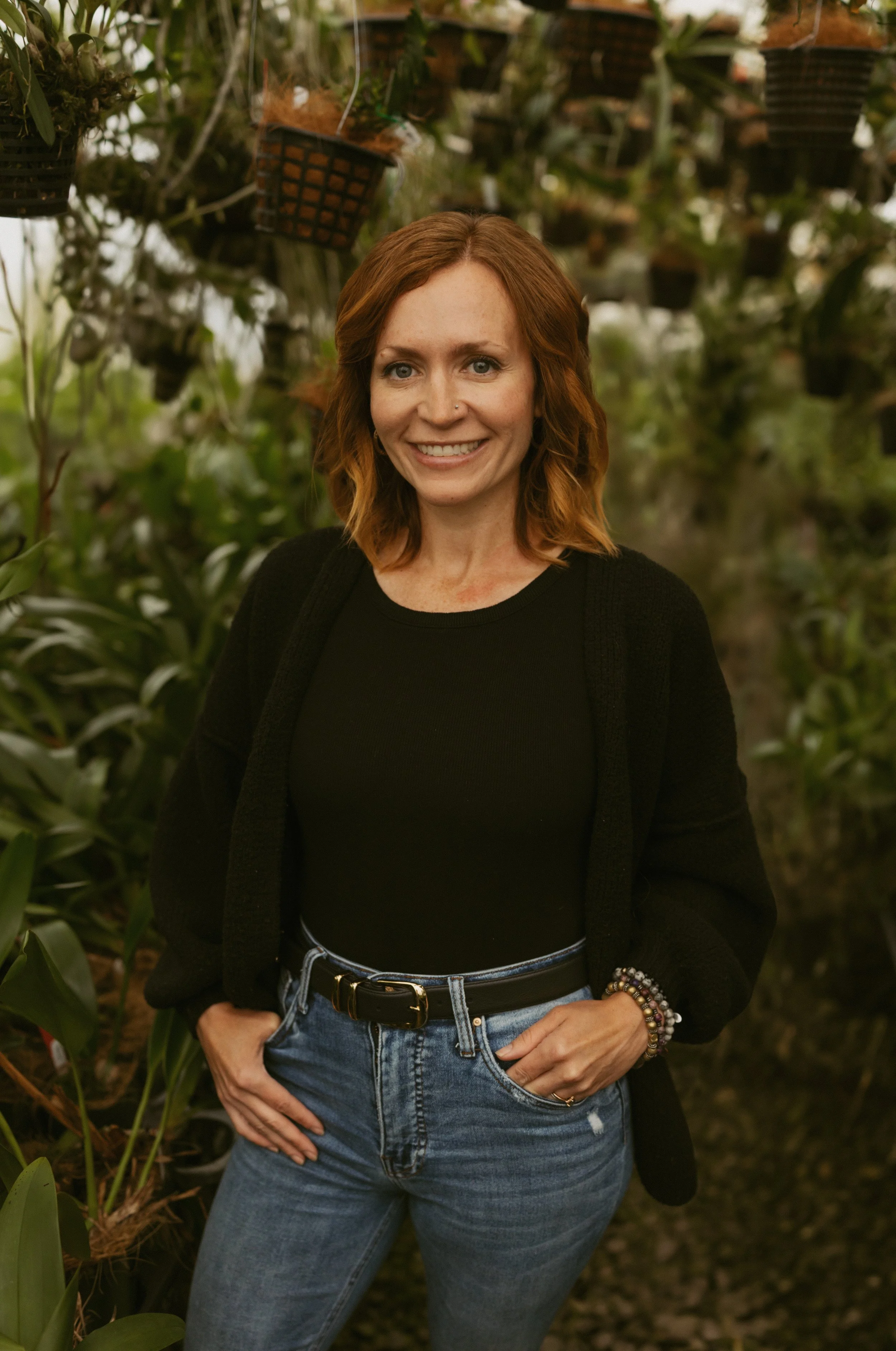 A woman with auburn hair, smiling, stands in a greenhouse filled with hanging pots of plants, wearing a black top, black cardigan, and blue jeans.