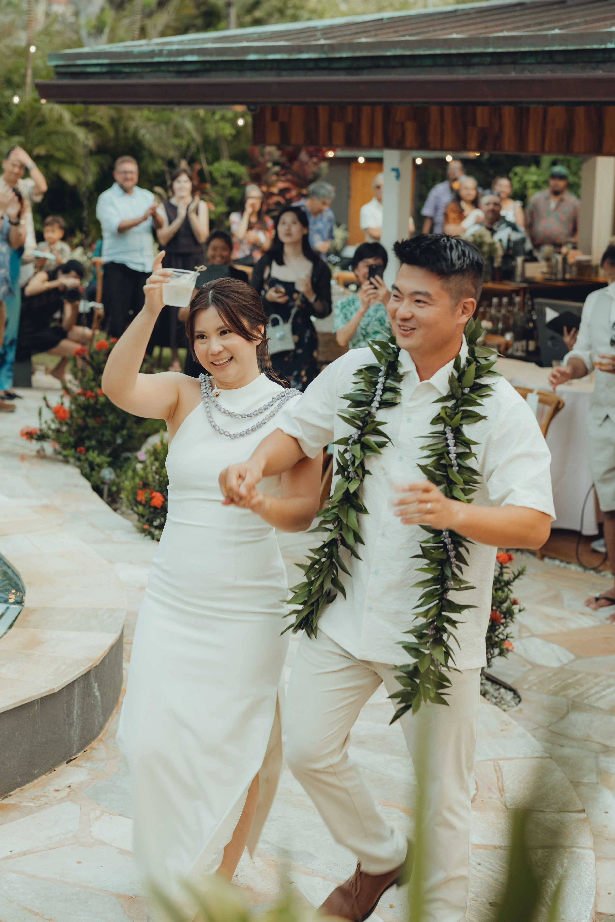 Emma and Evan celebrating in white wedding attire and lei, surrounded by their guests.