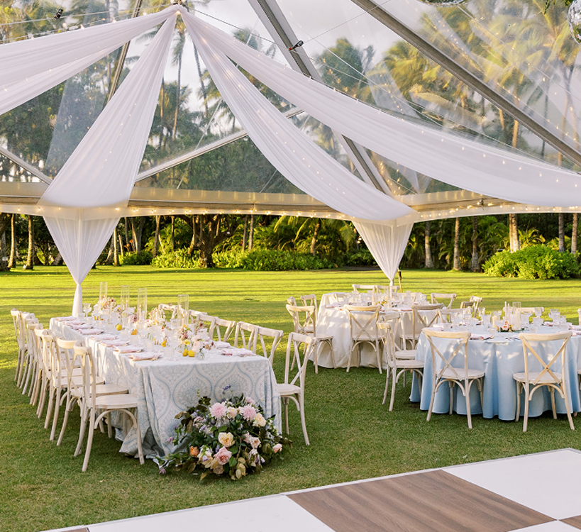 Outdoor wedding reception with white chairs, floral arrangements, and draped tent decor on a garden lawn.