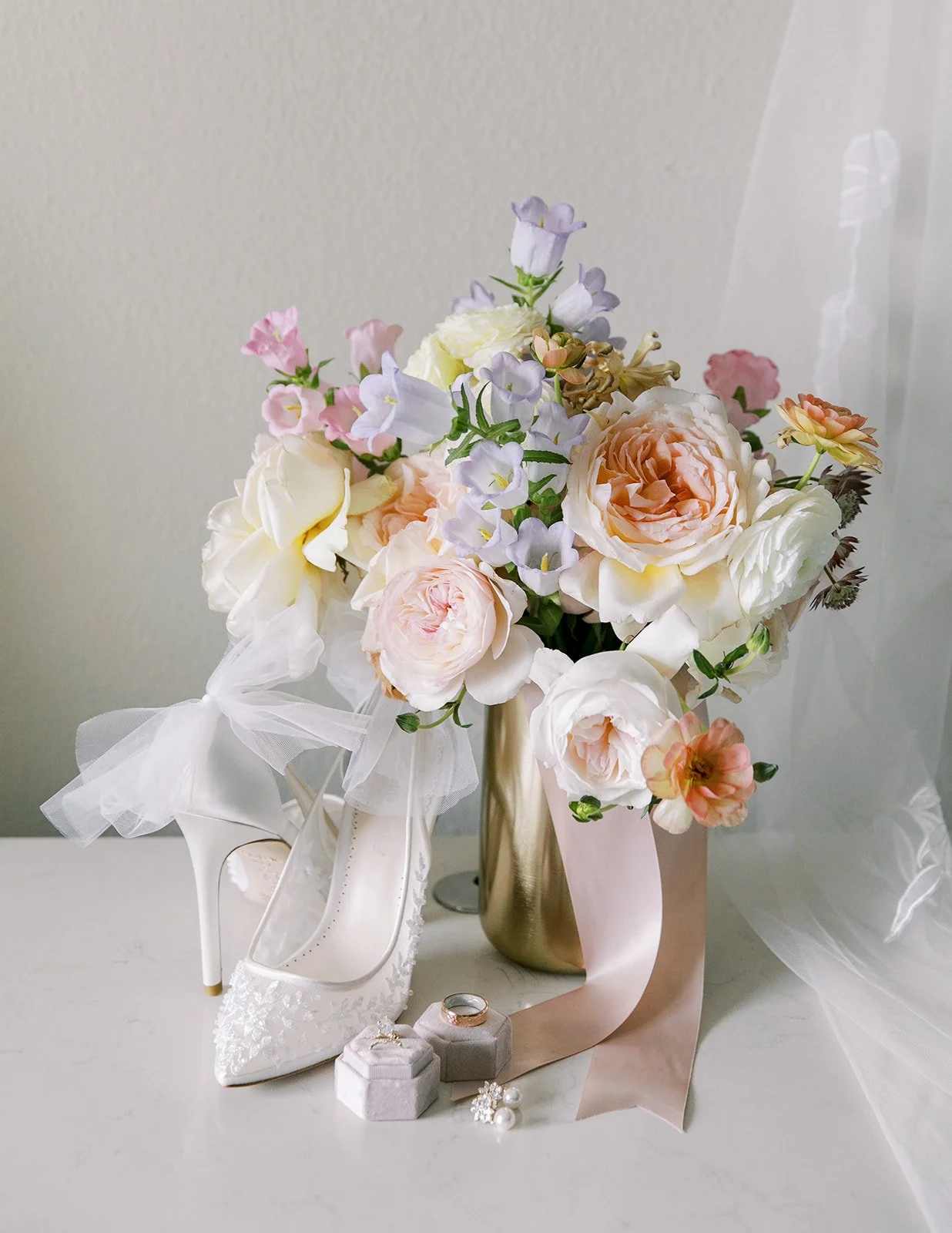 Bridal bouquet with pastel flowers displayed beside wedding shoes, rings, and soft fabric details.