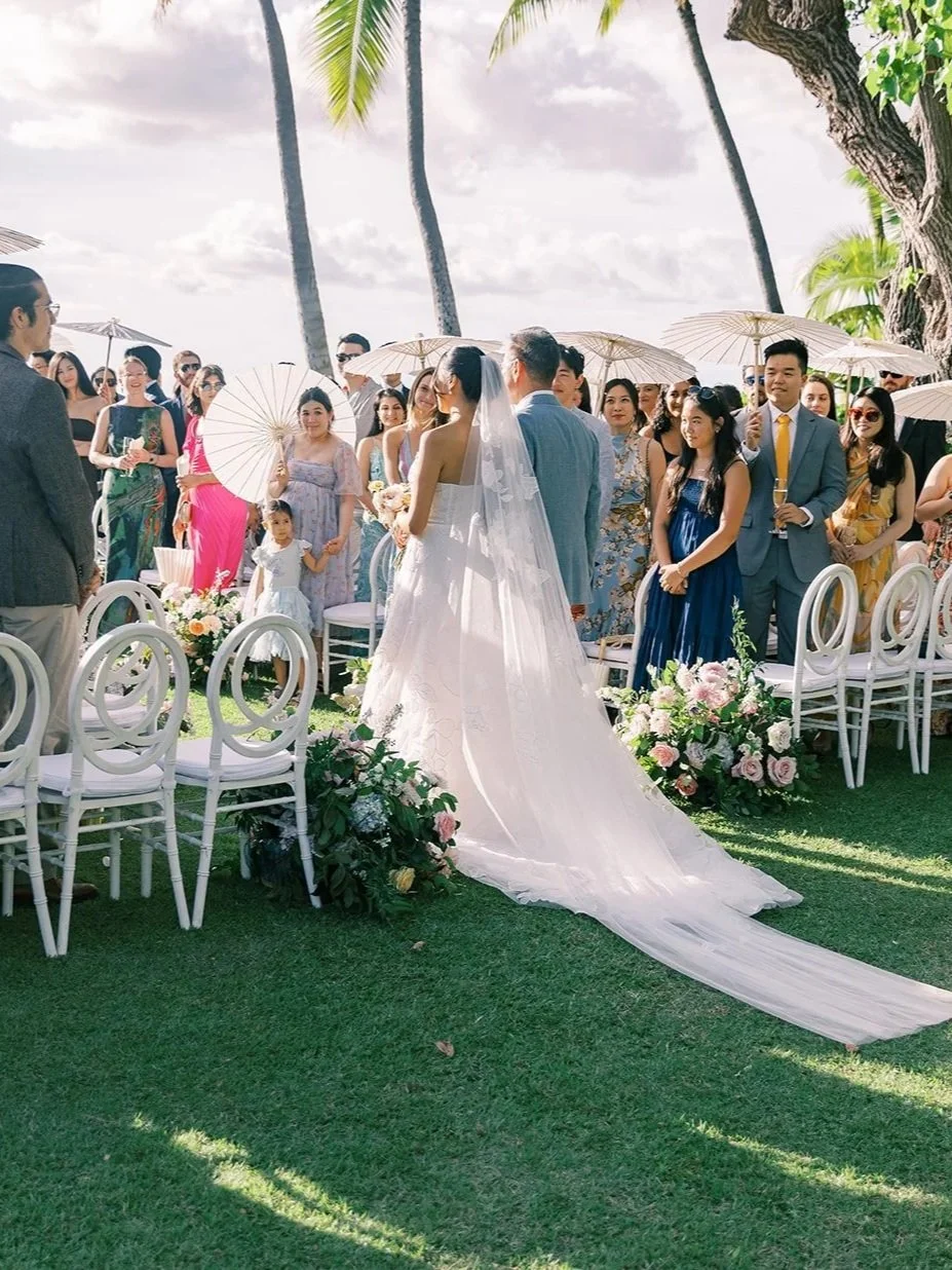 Bride walking down the aisle at an outdoor wedding ceremony surrounded by guests and floral arrangements.