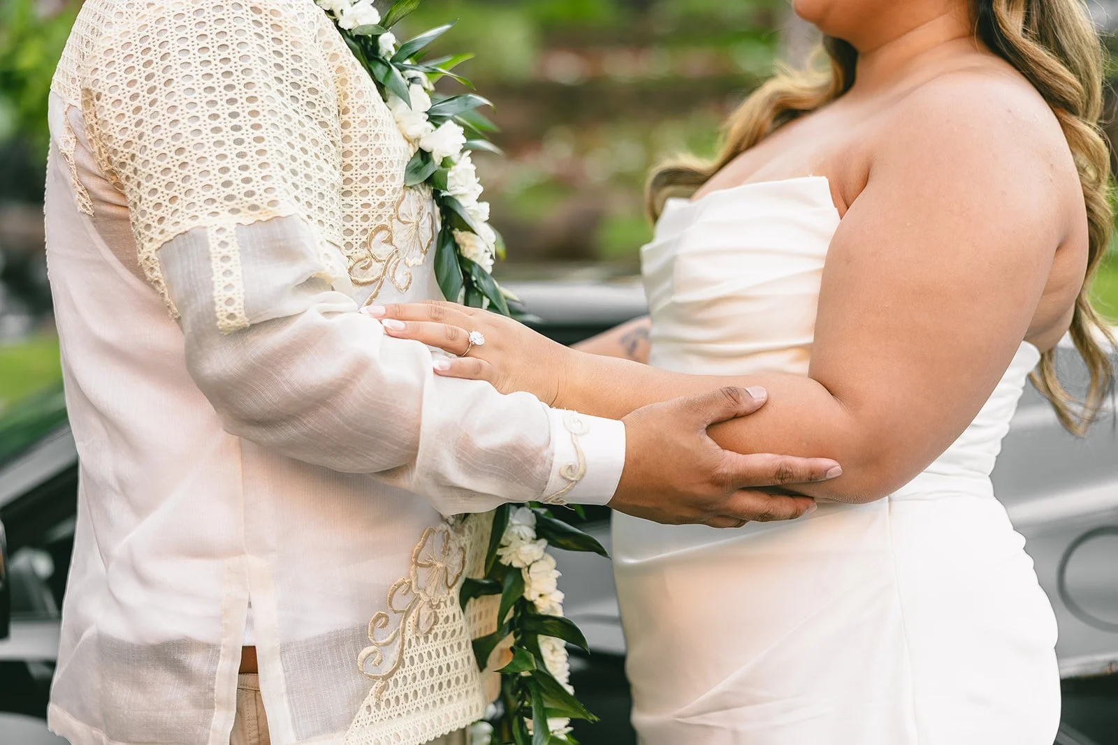 Close-up of a couple holding hands during a wedding ceremony, showing floral lei and wedding attire details.