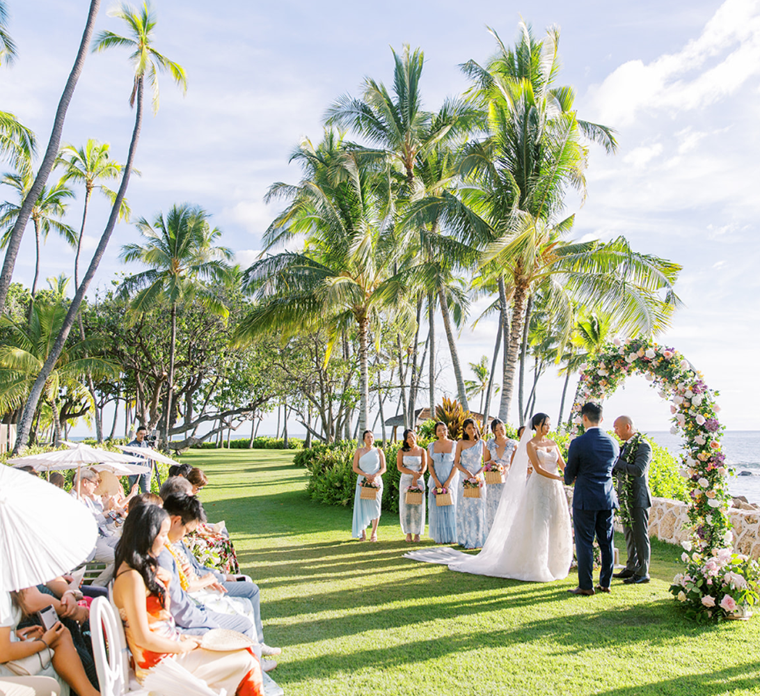 Outdoor wedding ceremony on a tropical lawn with the couple standing beside the bridal party under palm trees.
